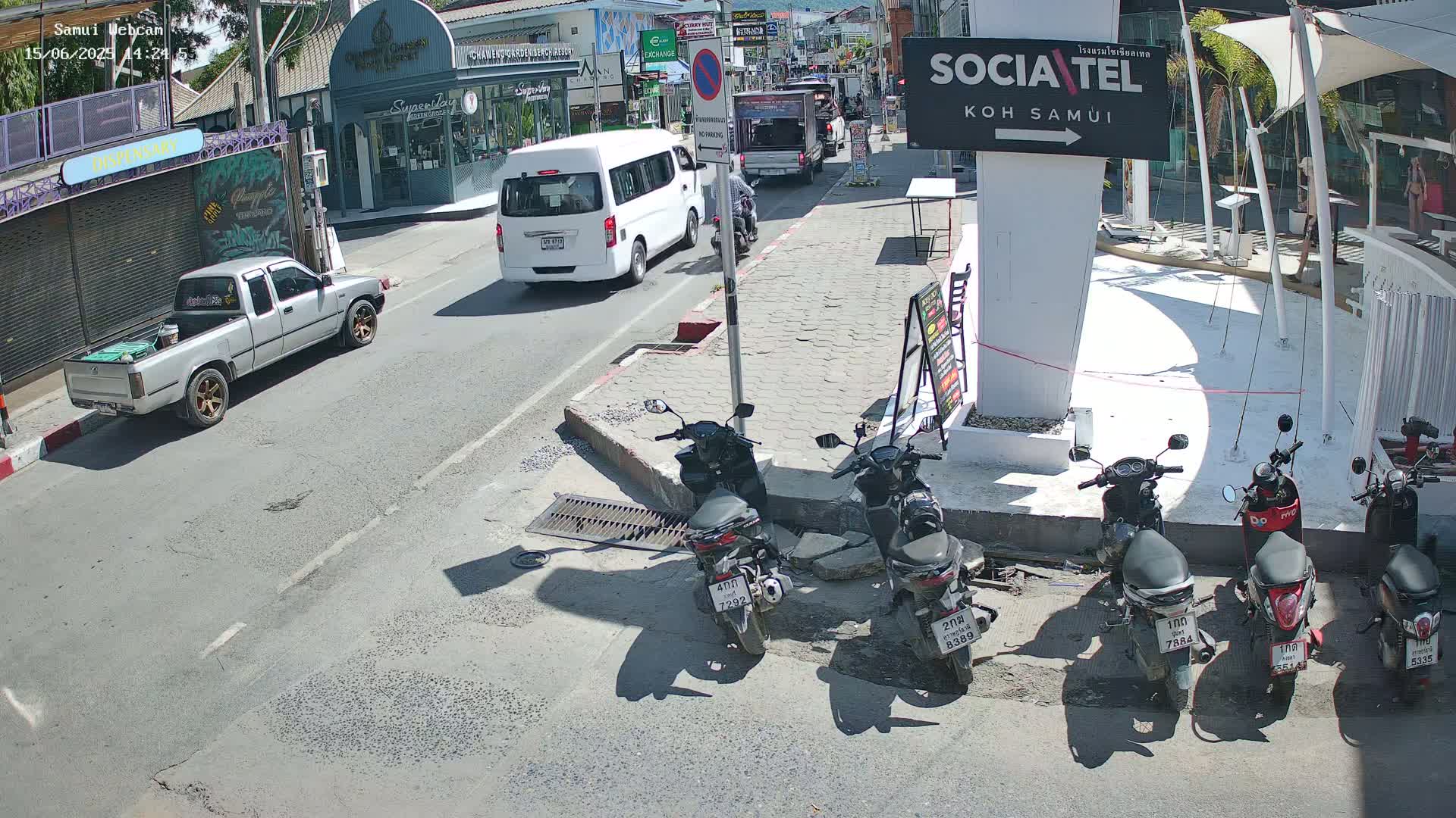 A sunny street scene shows several vehicles, including a pickup truck, van, and several parked motorbikes, alongside shops and buildings.