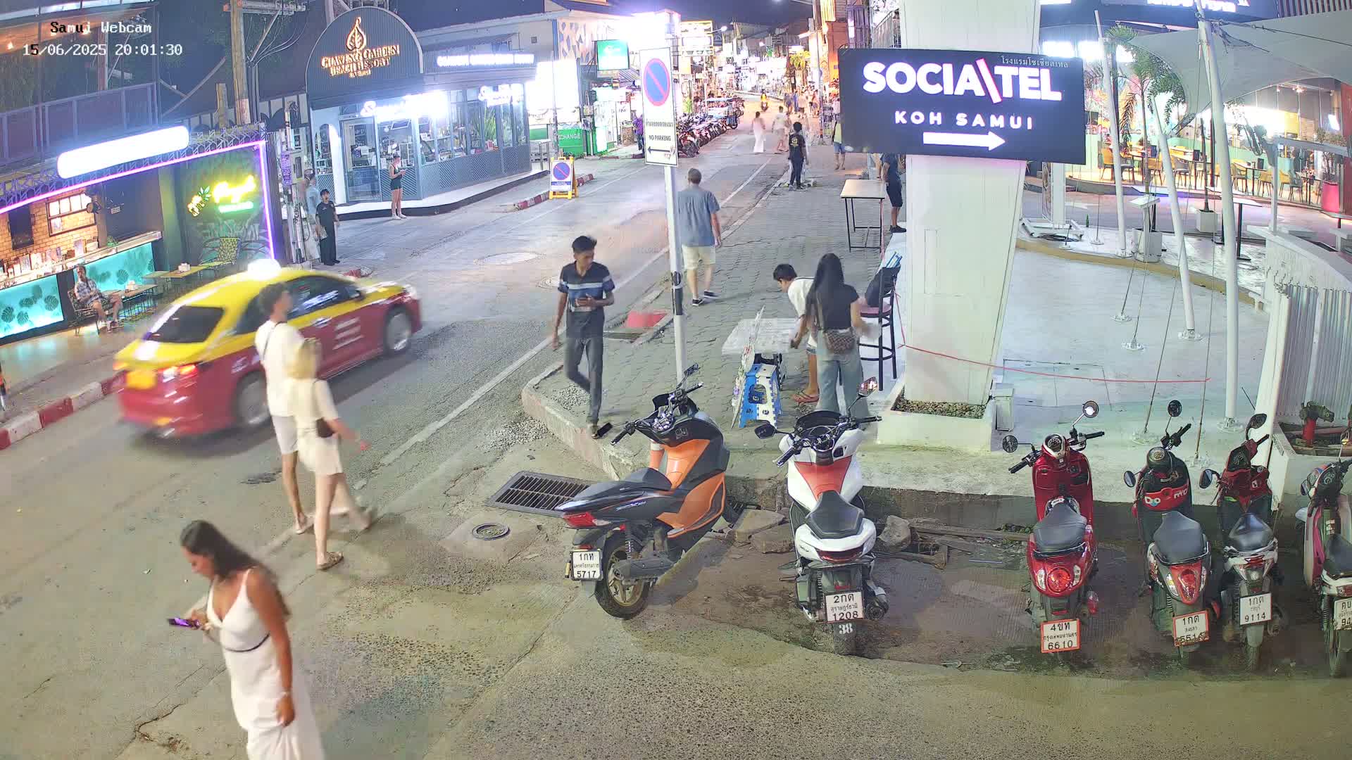 A nighttime street scene shows several pedestrians, a yellow taxi, and numerous parked motor scooters under clear skies.