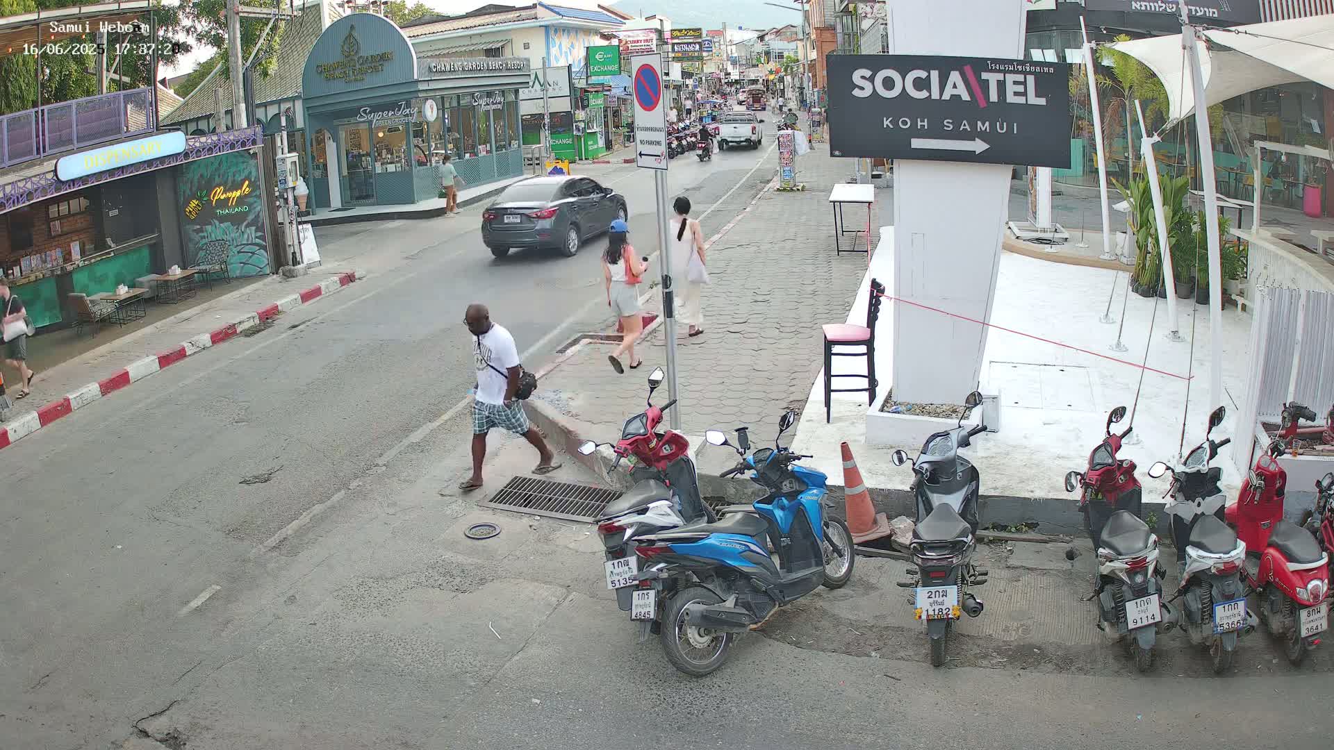 A street scene shows several pedestrians walking, a few cars driving, and numerous parked motorbikes along the side of a road lined with shops and restaurants under a partly sunny sky.