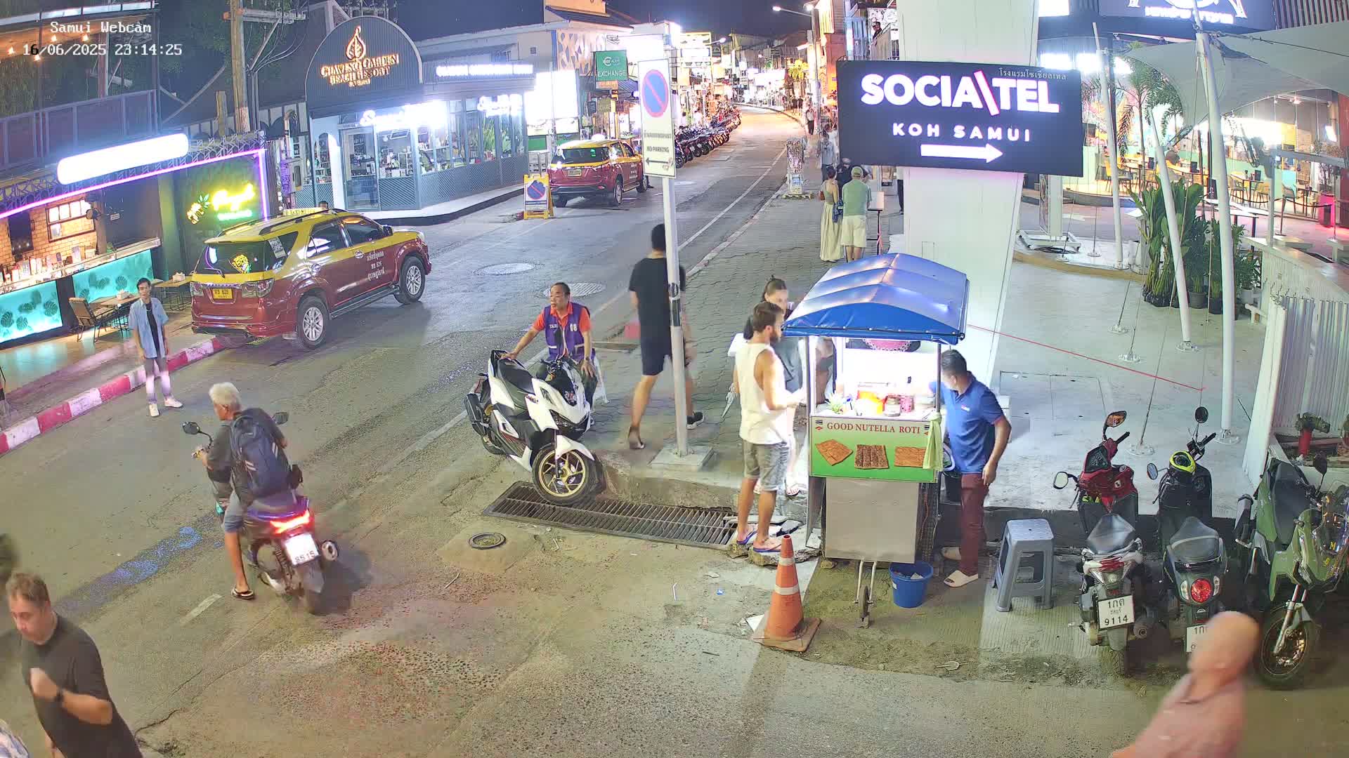 At night, several people walk and drive motorbikes on a street lined with shops and restaurants, and a food vendor stands by their cart.