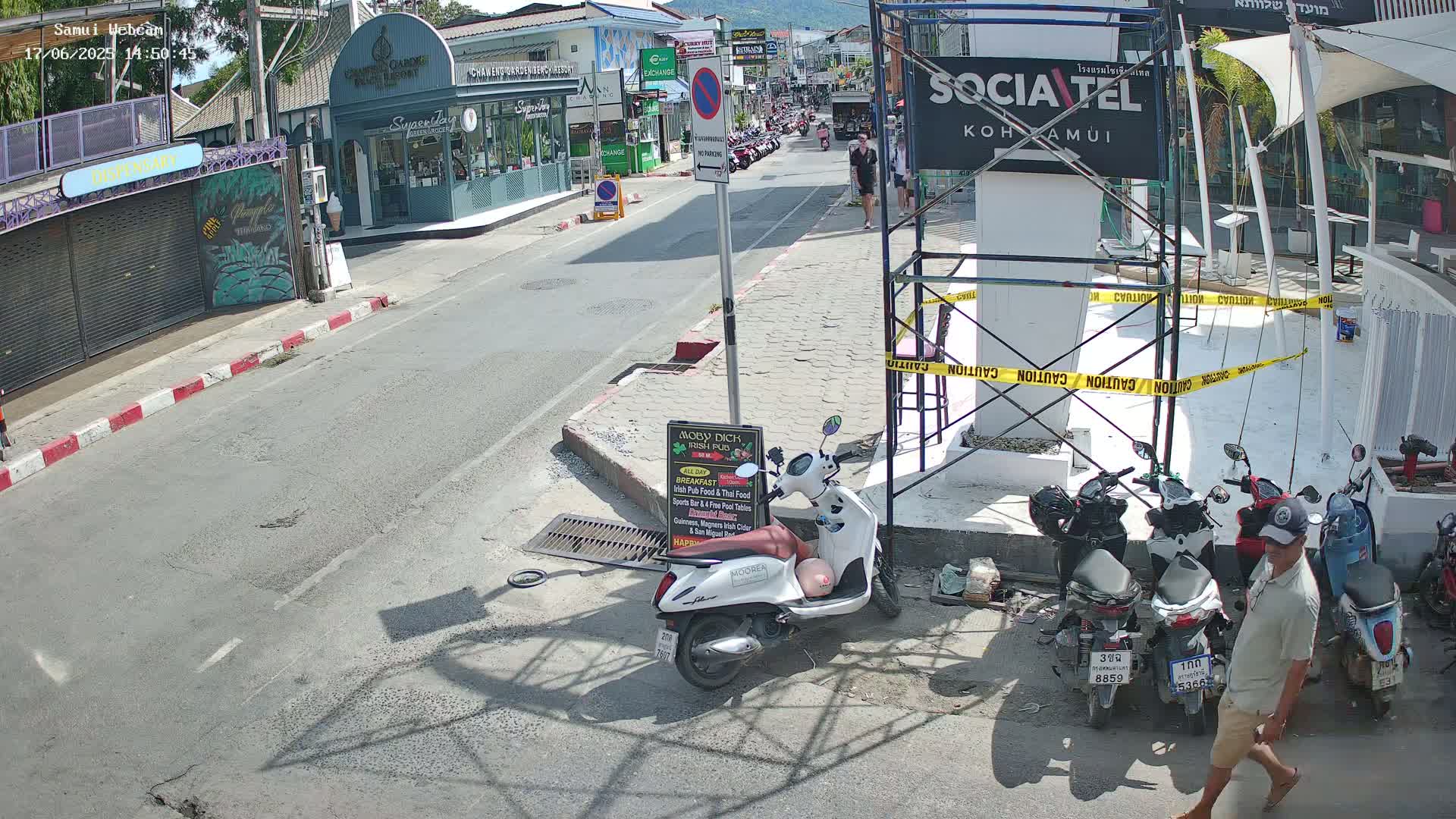 A sunny street scene shows a road with parked motorbikes and shops lining either side, construction scaffolding near a sign, and a man walking.