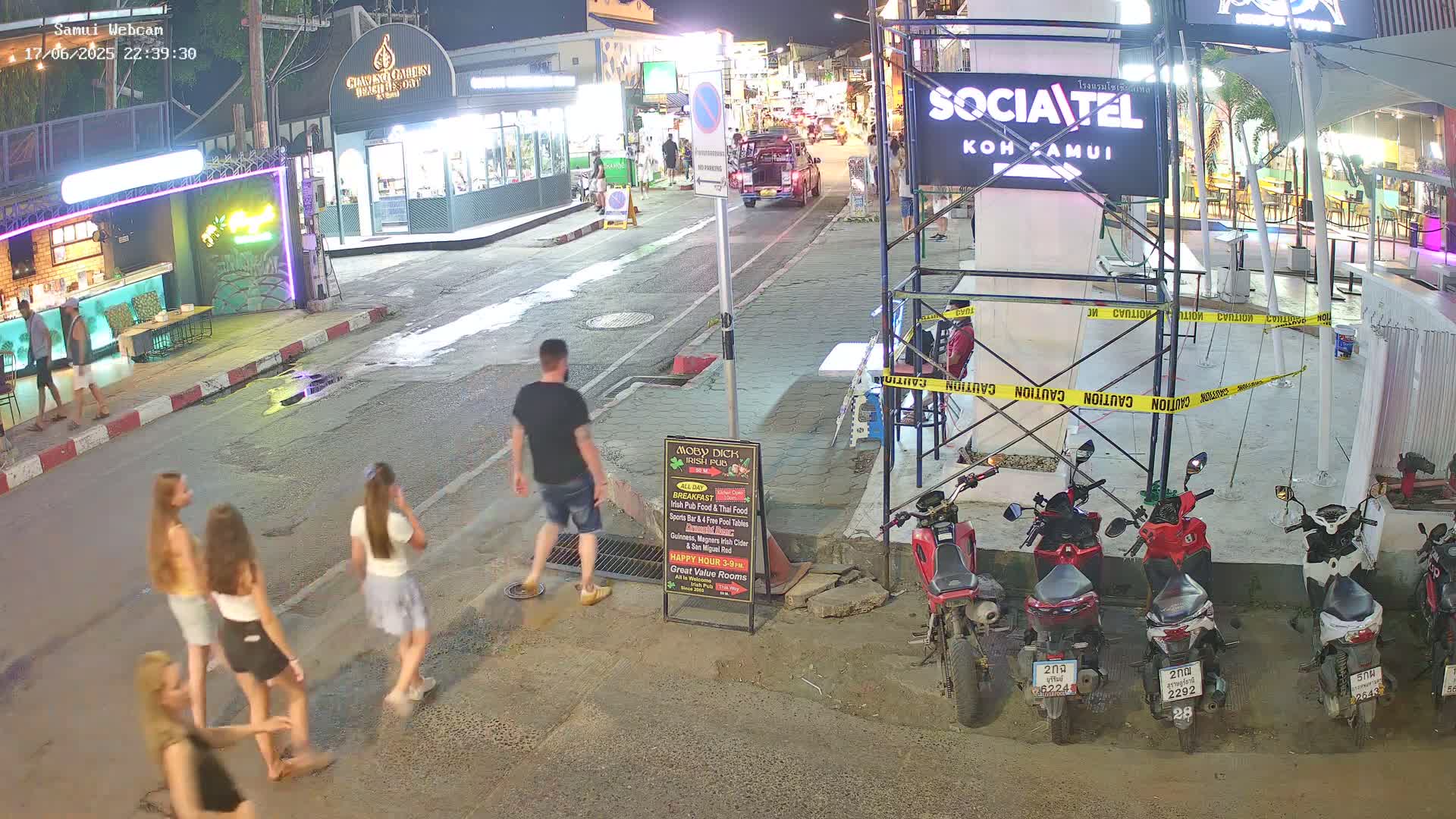 At night, several people walk along a street lined with buildings and parked motorbikes under construction scaffolding; some motorcyclists are near the construction.