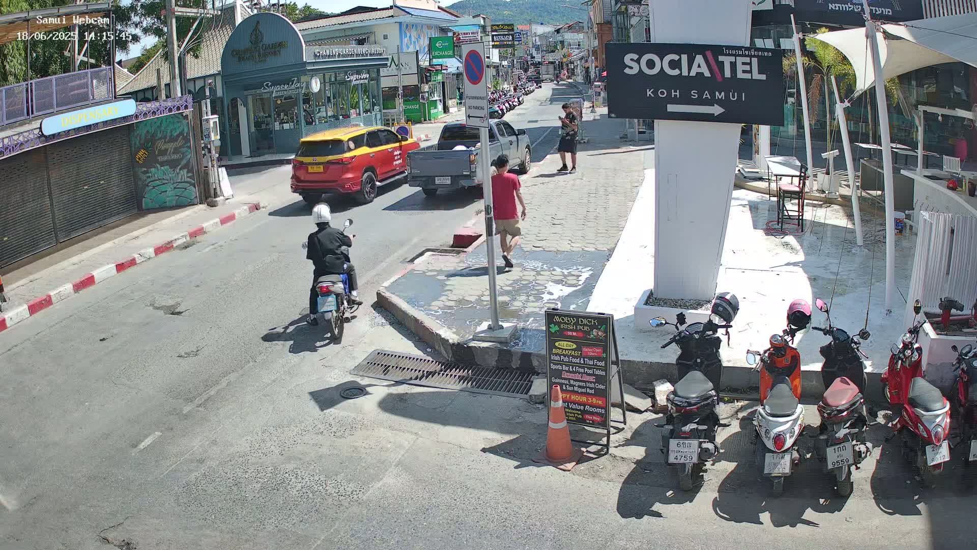 A sunny street scene shows several parked motorbikes and cars, with pedestrians and a motorcyclist on the road.