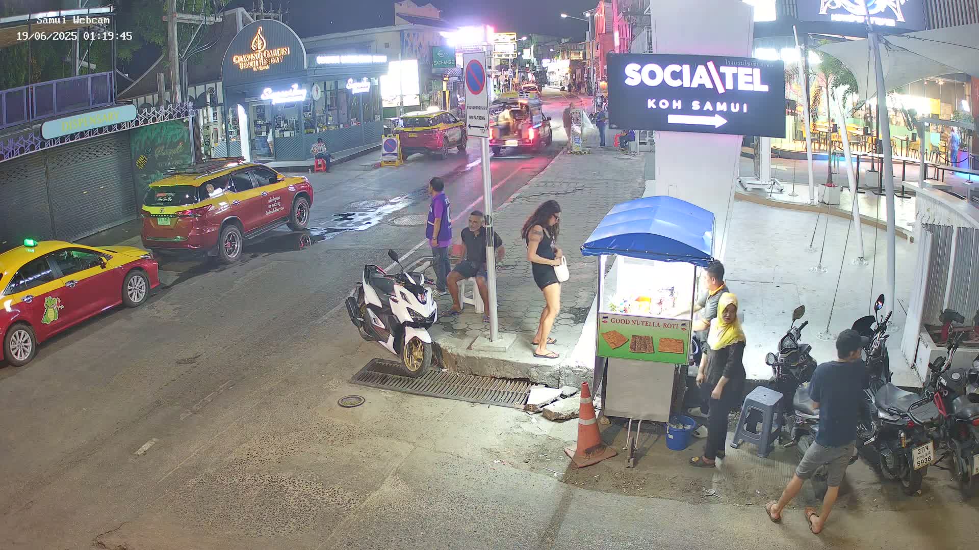 At night, on a city street with parked cars and several people standing around a food stall, a few motorcycles are parked nearby.