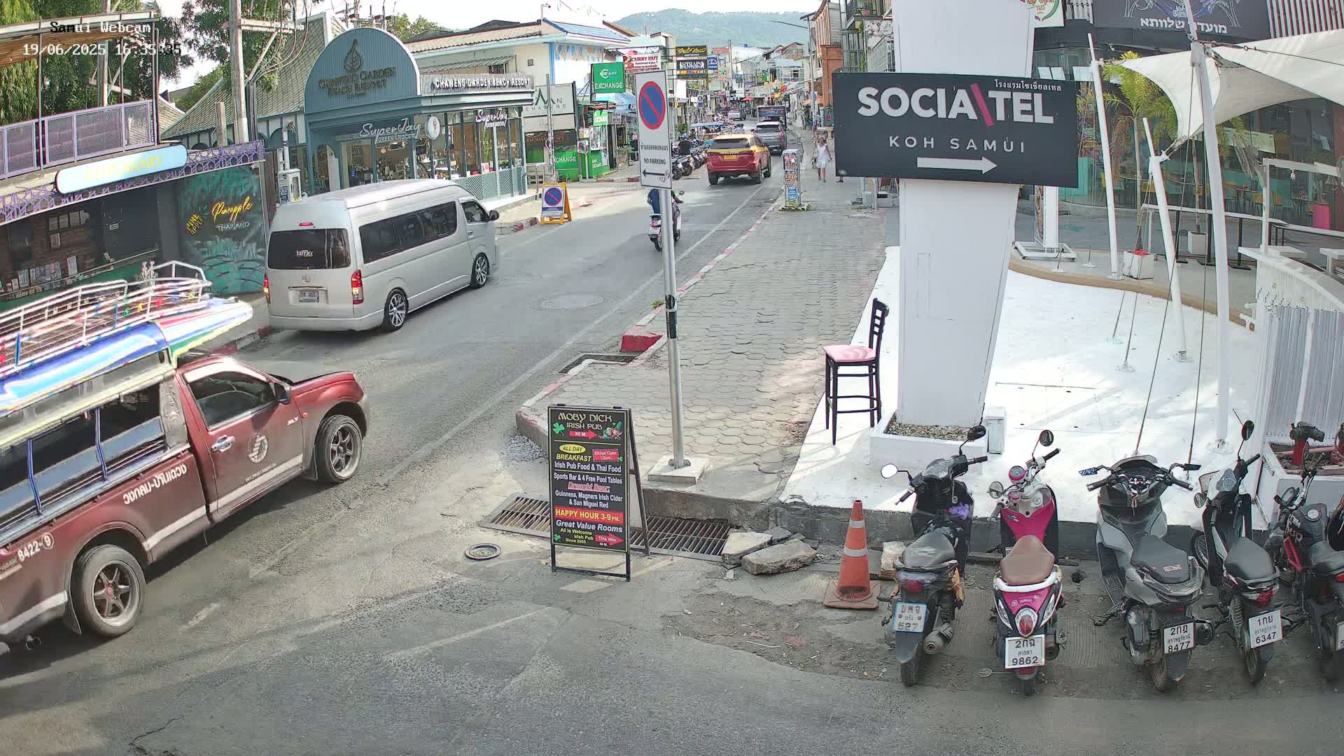A sunny street scene shows a red pickup truck and a silver van parked on the side of the road, with several motorbikes parked along the sidewalk and other vehicles driving down the street.