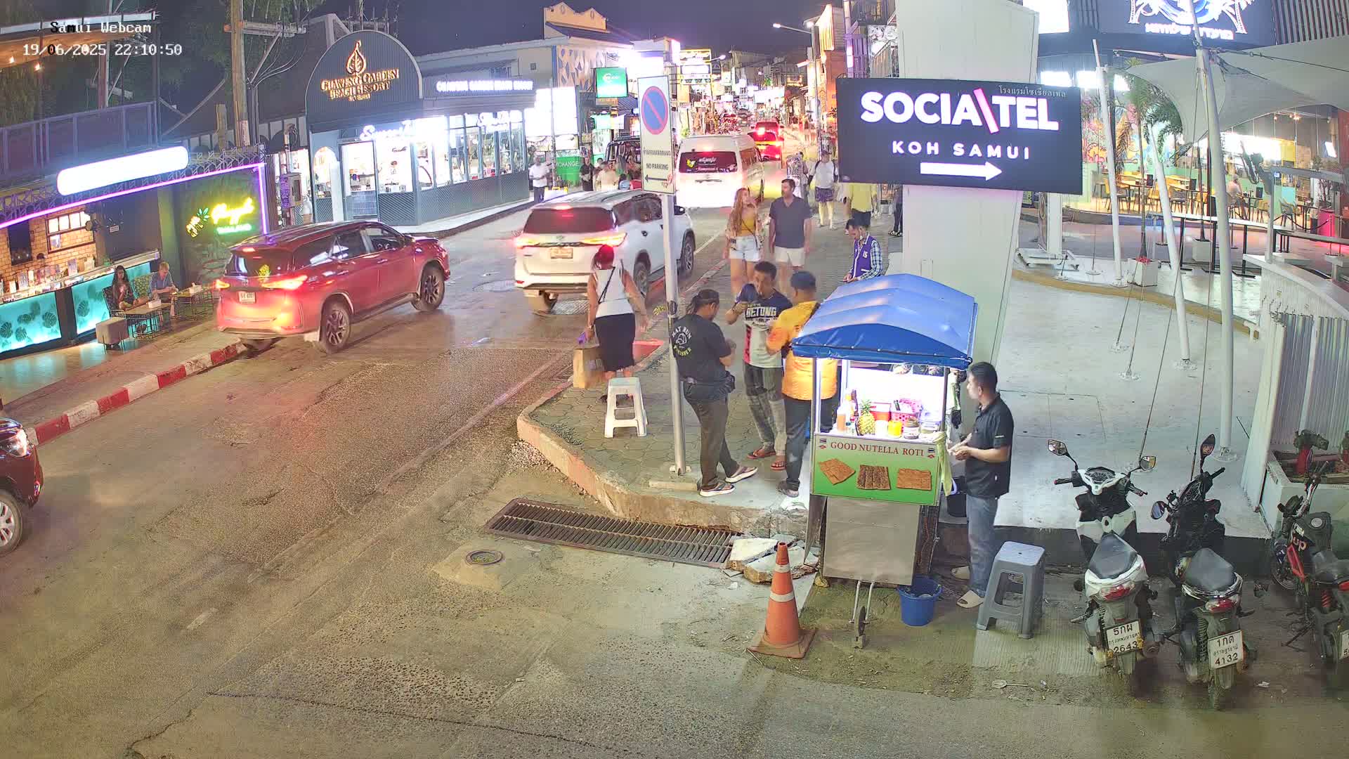 At night, several people stand near a food cart and parked cars on a street with some pedestrian traffic.