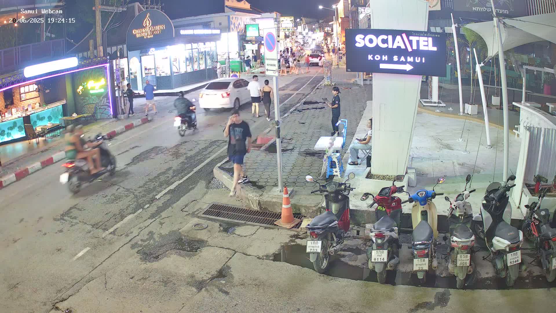 A nighttime street scene shows pedestrians and motorcyclists on a road lined with shops and parked motorbikes, under partly cloudy skies.
