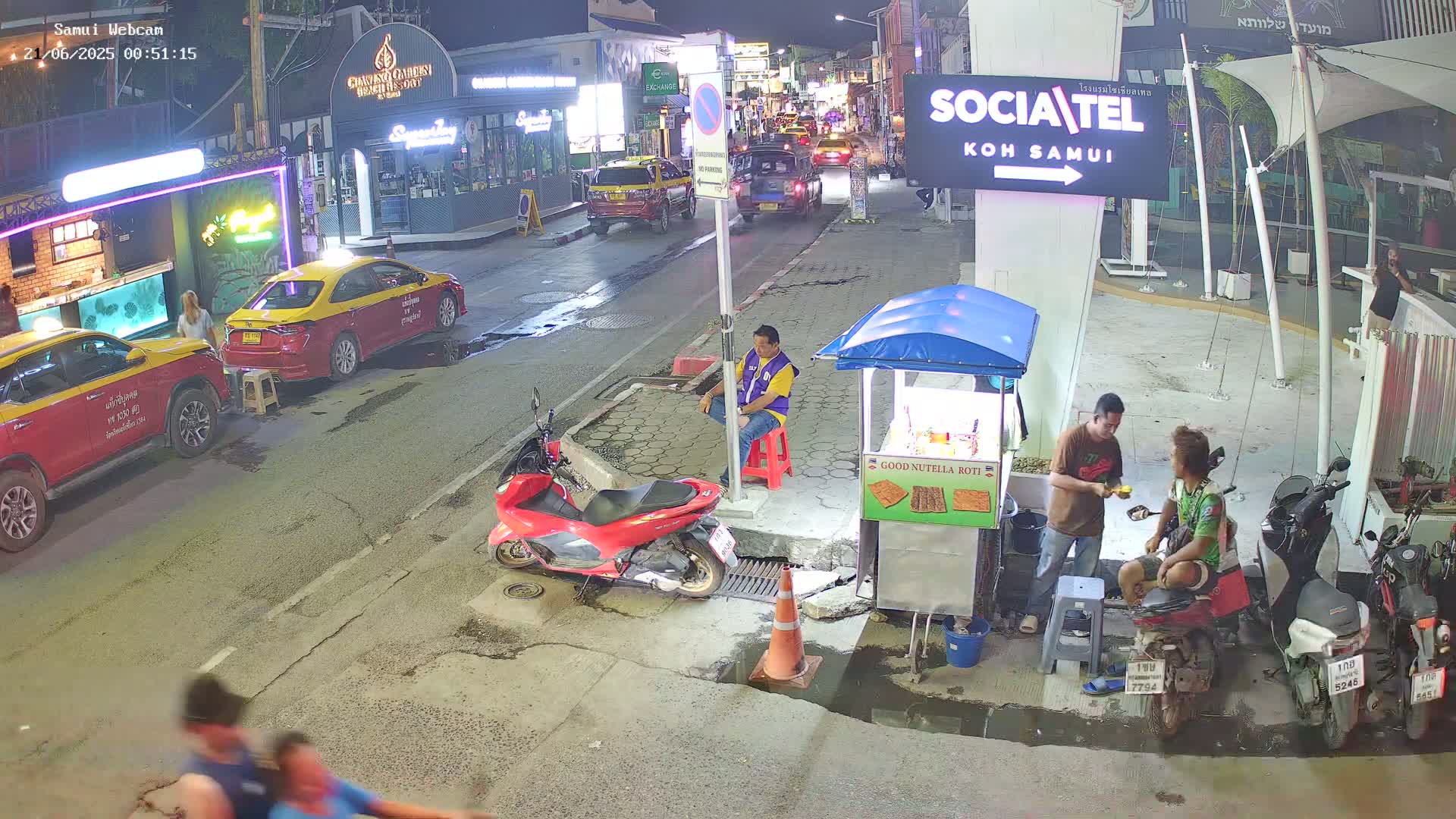 A nighttime street scene shows parked cars, motorbikes, and a few people, including one sitting at a small food stand, with puddles on the wet pavement.