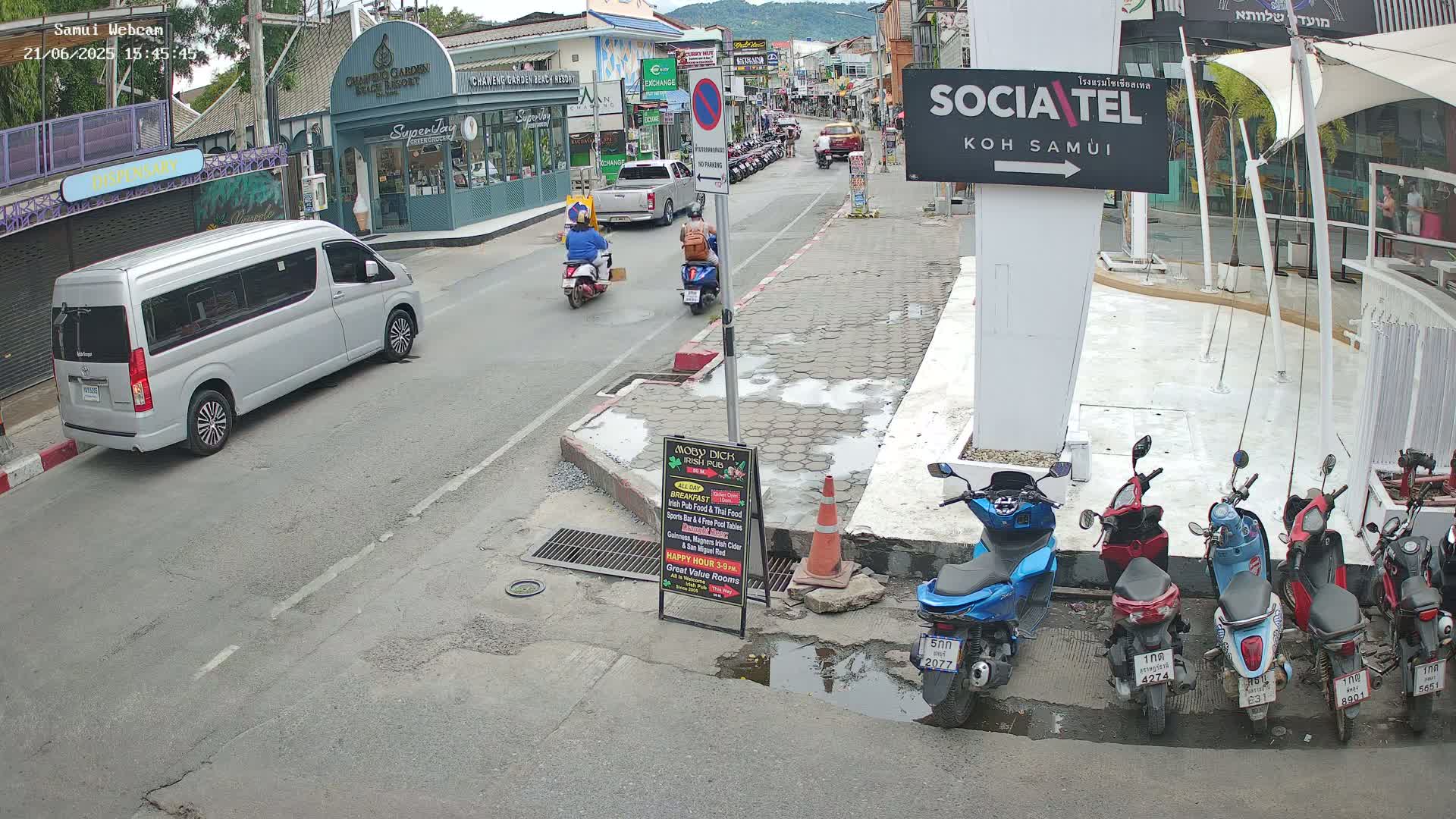 A silver minivan is parked on a street in a town, with several mopeds parked nearby and two people riding mopeds down the street, under partly sunny skies.