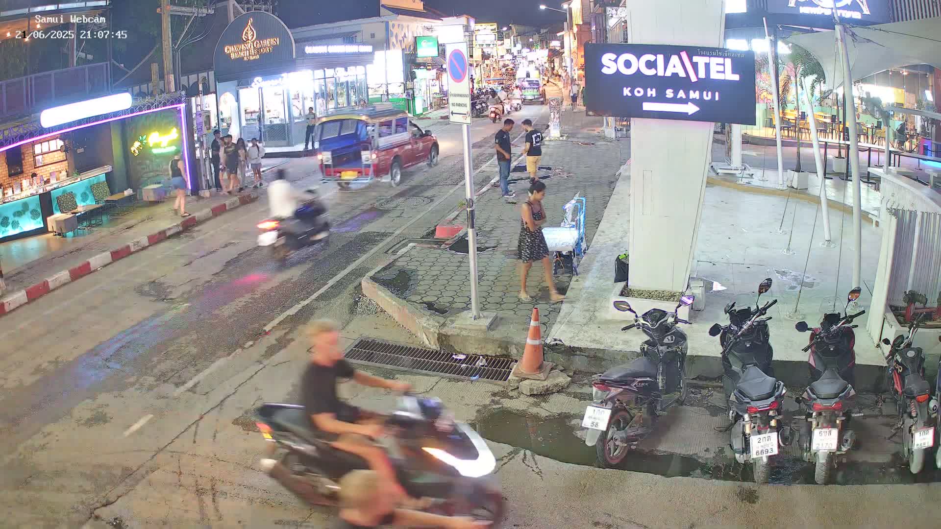 A nighttime street scene shows people walking and riding motorbikes past shops and restaurants, with several parked motorbikes along the side of the road.