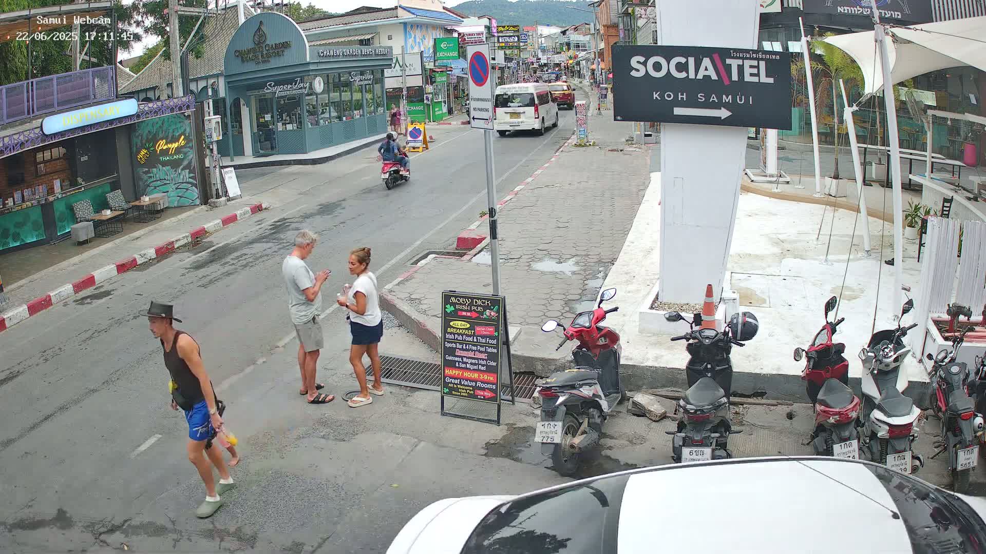 A street scene shows several people standing on a city street, some motorbikes parked along the sidewalk, and a few vehicles driving down the road in bright daylight.