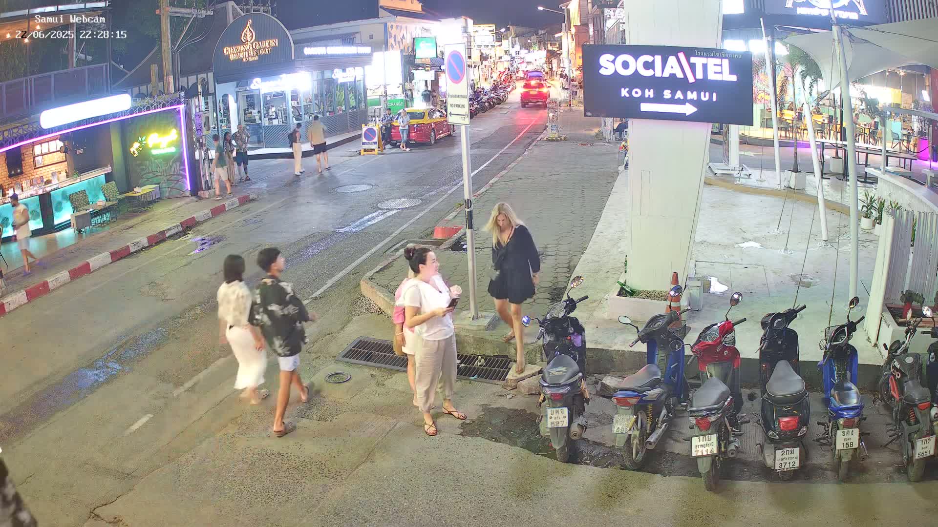 At night, several people walk along a street lined with shops and parked motorbikes under clear skies.