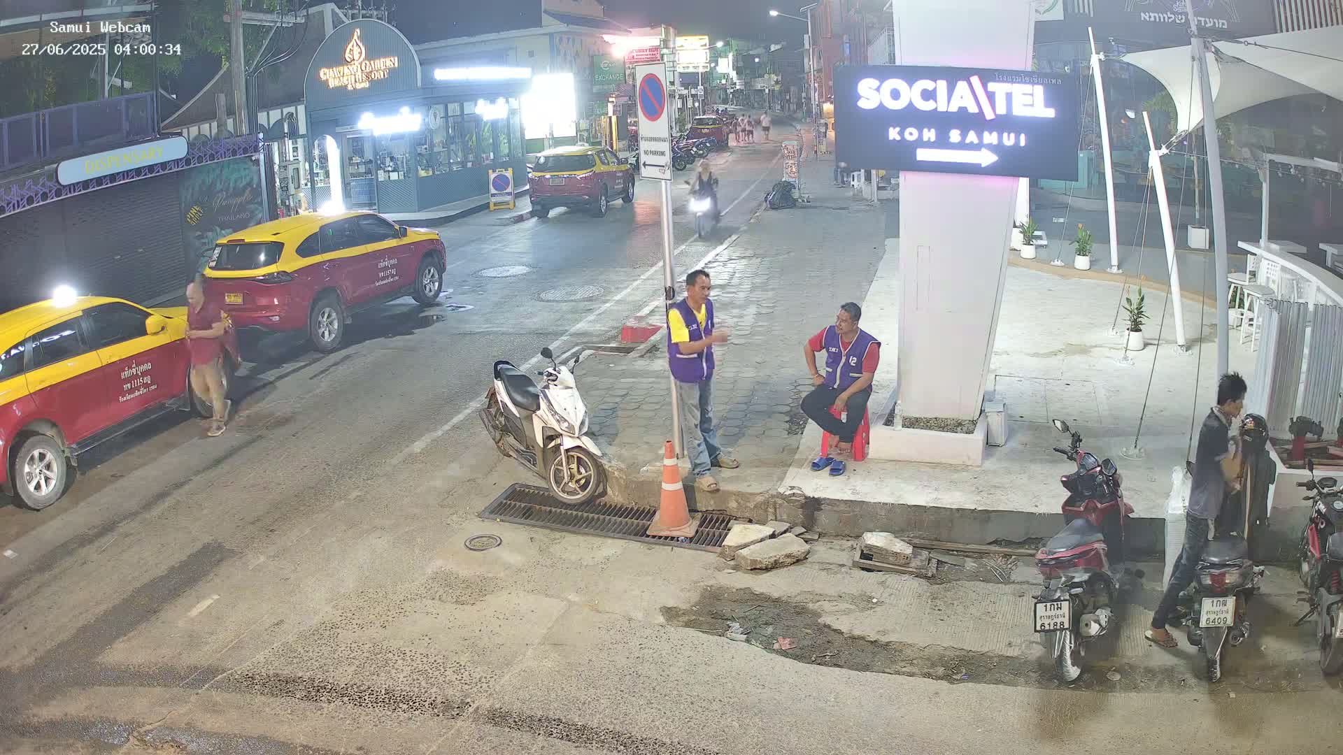 At night, several people stand near parked taxis and motorcycles on a street with damaged pavement, under clear skies.