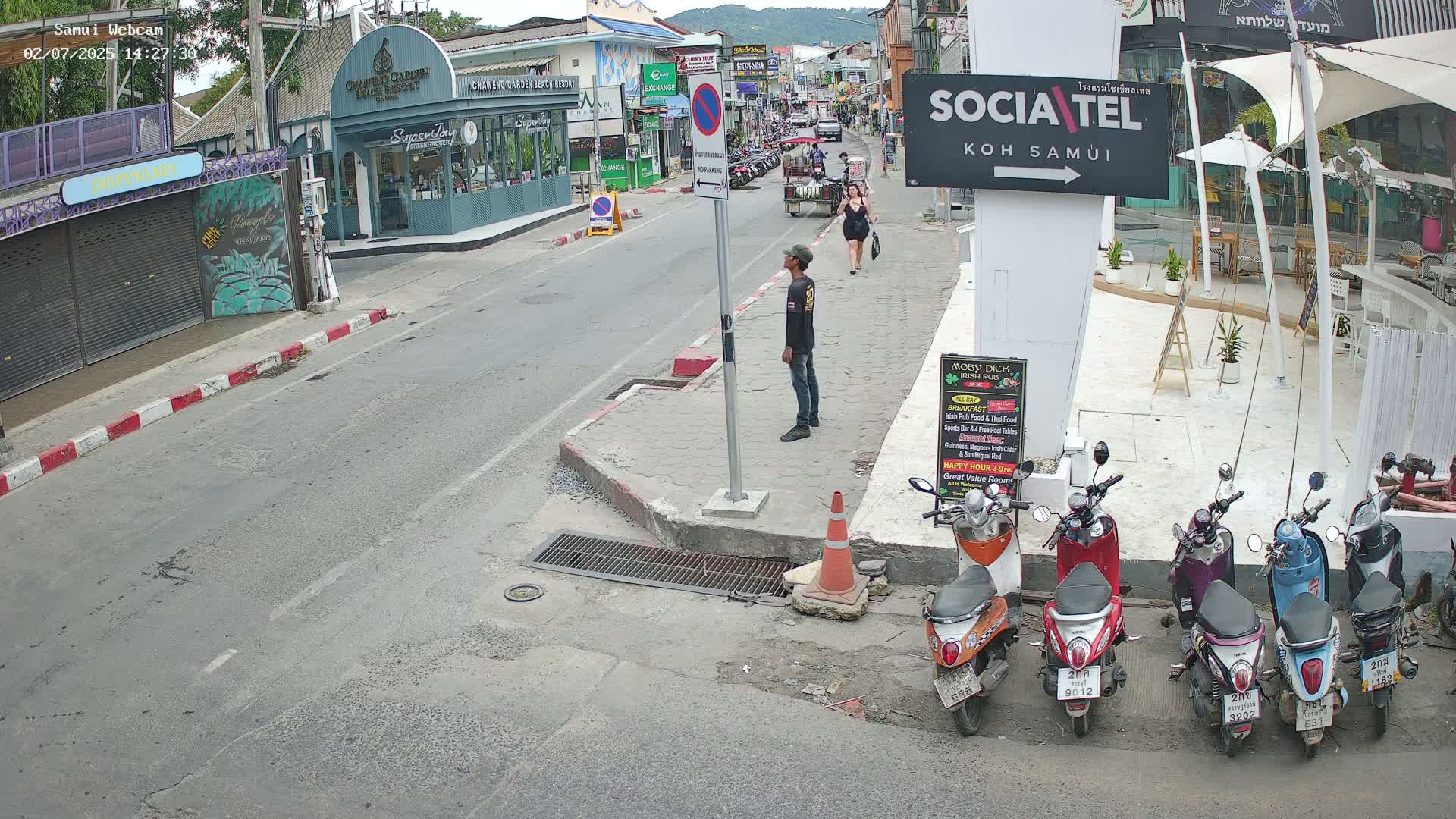 A street scene on a sunny day shows a man standing on a sidewalk next to a row of parked motor scooters, with a woman walking further down the street near shops and restaurants.