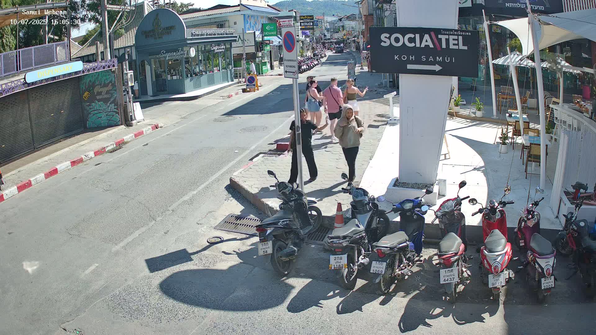 A sunny street scene shows several people walking among parked motor scooters in front of shops and restaurants.