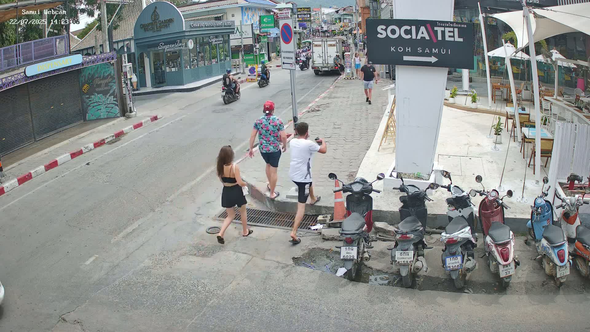 Three people walk on a sidewalk past parked motorbikes near a street with cars and buildings in sunny weather.
