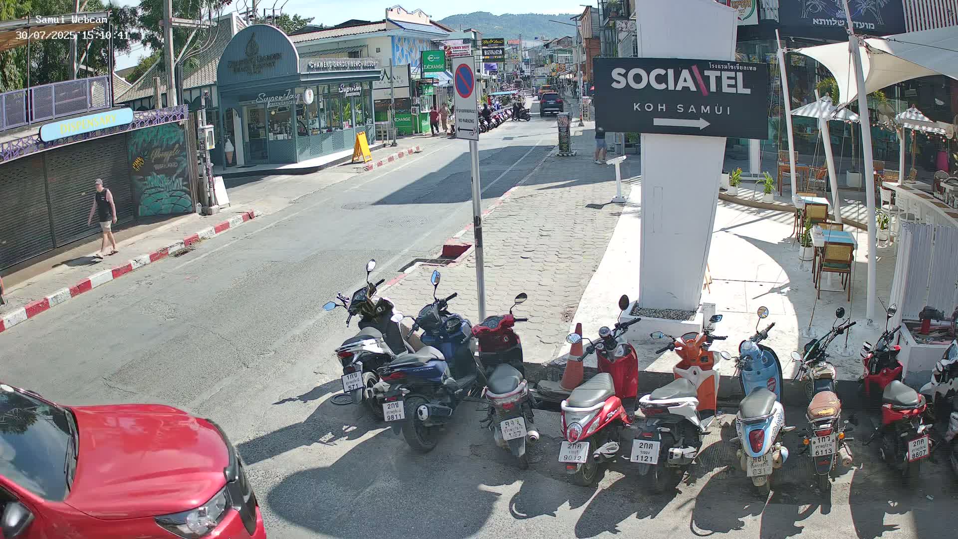 A sunny street scene shows a red car in the foreground, several parked motorbikes, and a street lined with shops and restaurants.