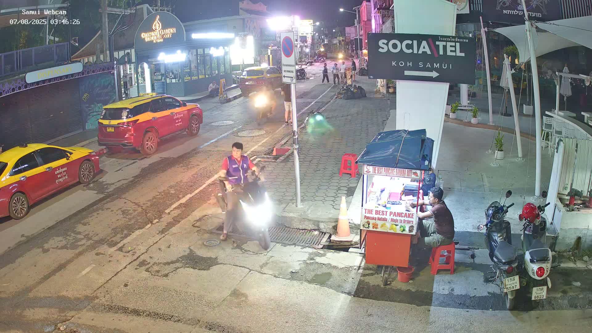 A nighttime street scene shows a person on a motor scooter near a food cart, parked cars, and a few pedestrians, under well-lit conditions.