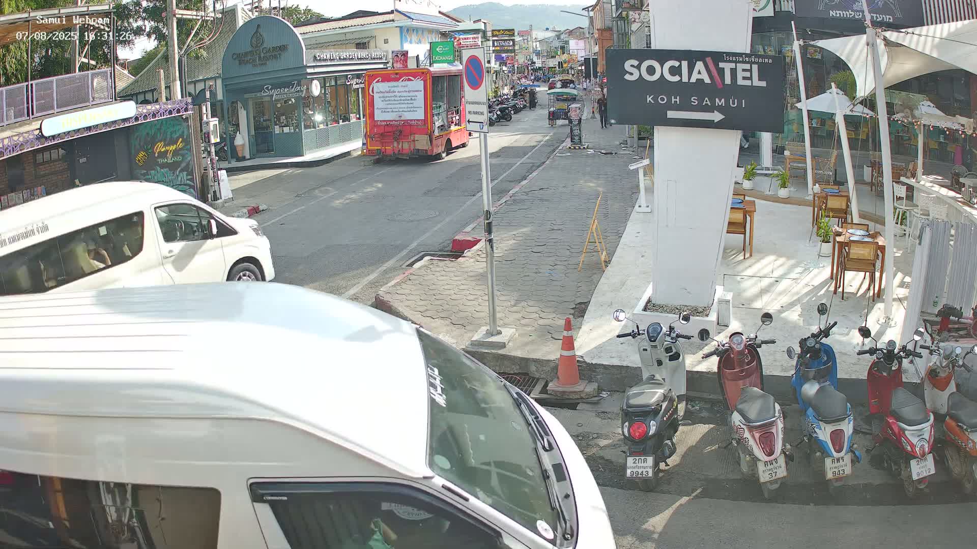 A sunny street scene shows several parked motorbikes next to a restaurant, with vans and a Coca-Cola truck visible on a road lined with shops and buildings.
