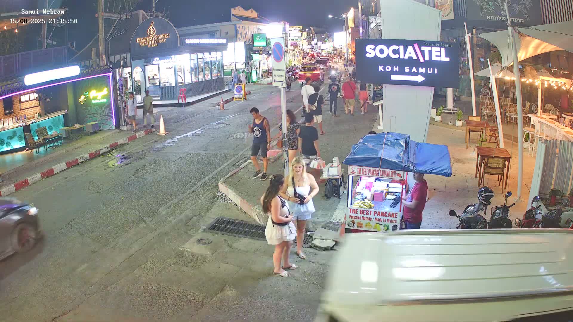 A sunny street scene shows several parked motorbikes next to a restaurant, with vans and a Coca-Cola truck visible on a road lined with shops and buildings.