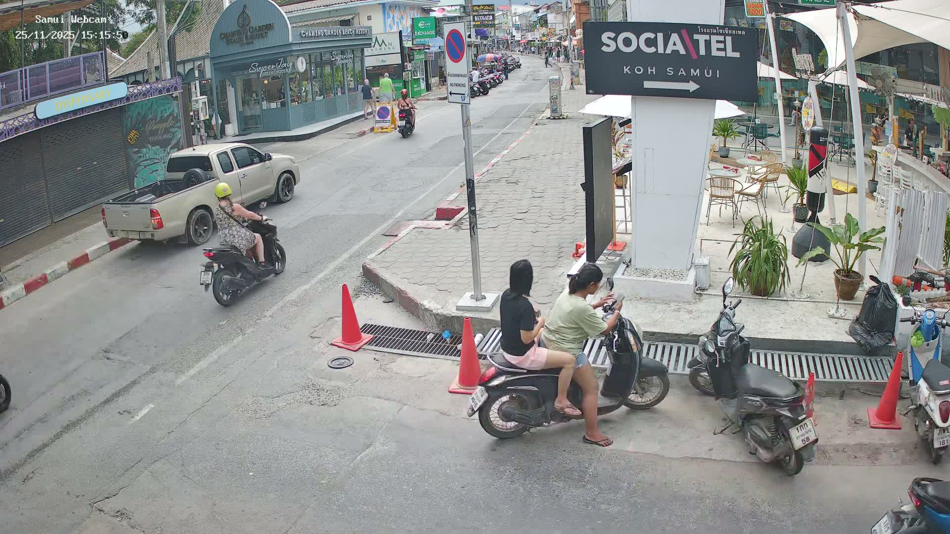 On a bright day, a busy street scene unfolds with multiple motorcycles, including two women sitting on one in the foreground and another rider passing a parked pickup truck, while pedestrians and storefronts line the road in what appears to be a commercial district.