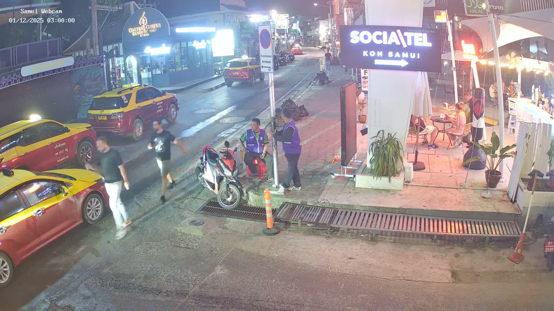 A sunny street scene in what appears to be a town, featuring a person walking on a sidewalk, parked motorbikes, and storefronts, with patches of water on the ground under bright sunlight.