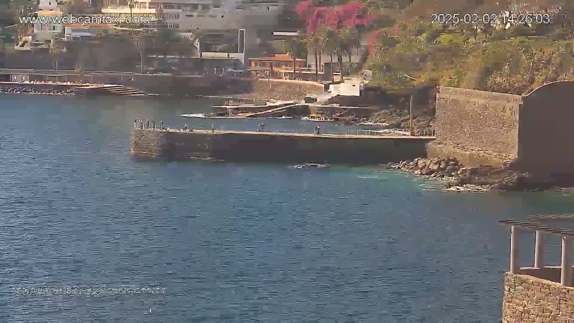 A stone pier extends into a calm, blue body of water next to a rocky shoreline with buildings and vegetation visible in the background under a sunny sky.
