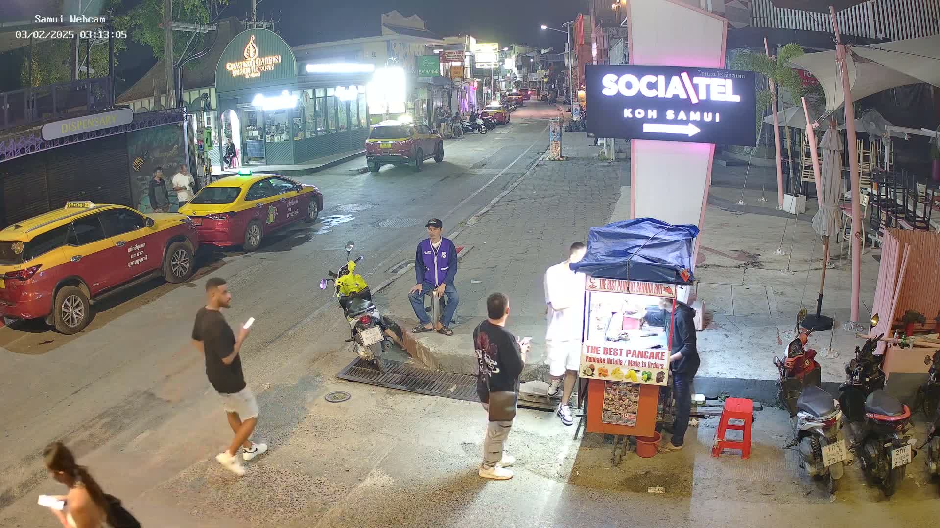 At night, several people stand and walk on a street in front of shops and food stalls, with parked cars and motorcycles nearby.