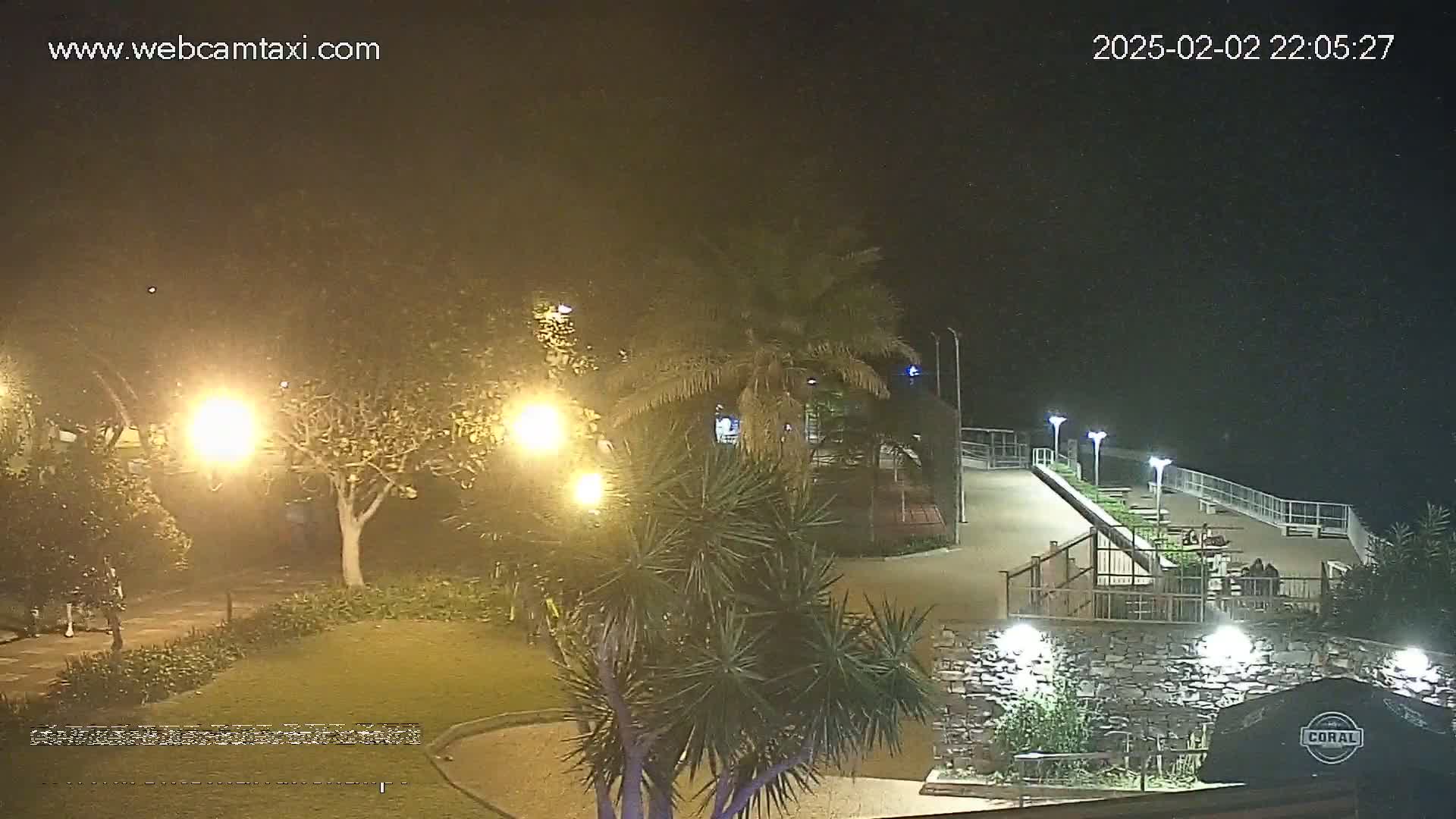 A nighttime view of a park-like area with palm trees and lights, overlooking a paved walkway and railing by a body of water under a slightly hazy sky.