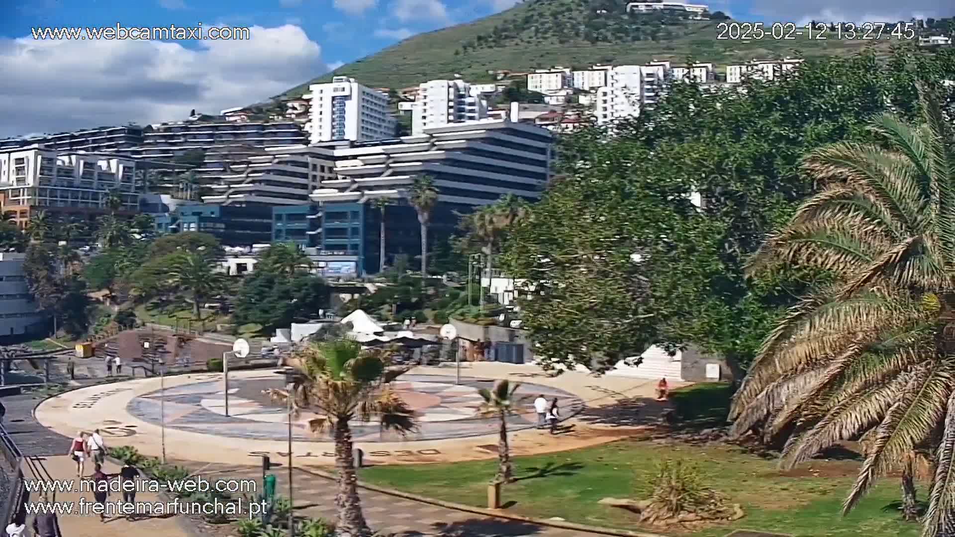 A sunny day overlooking a coastal town with multiple buildings, palm trees, and a paved plaza with people walking.
