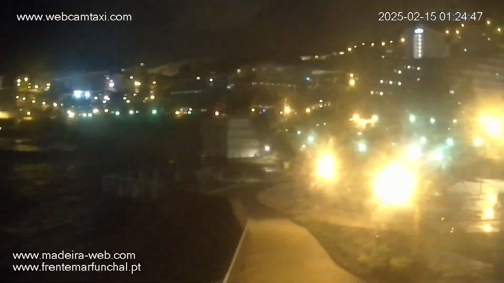 A nighttime view of a town illuminated by numerous lights, showing a coastal area with a path leading toward a brightly lit waterfront.