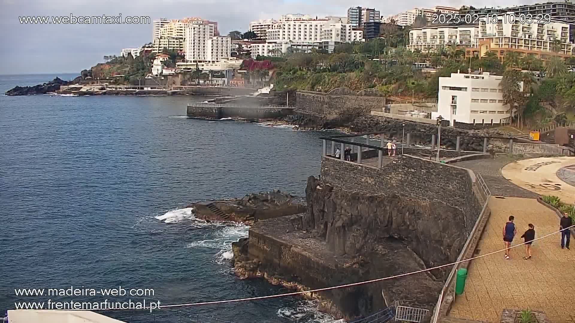 A coastal scene shows a rocky shoreline with a paved walkway, several buildings, and people walking, under overcast skies.