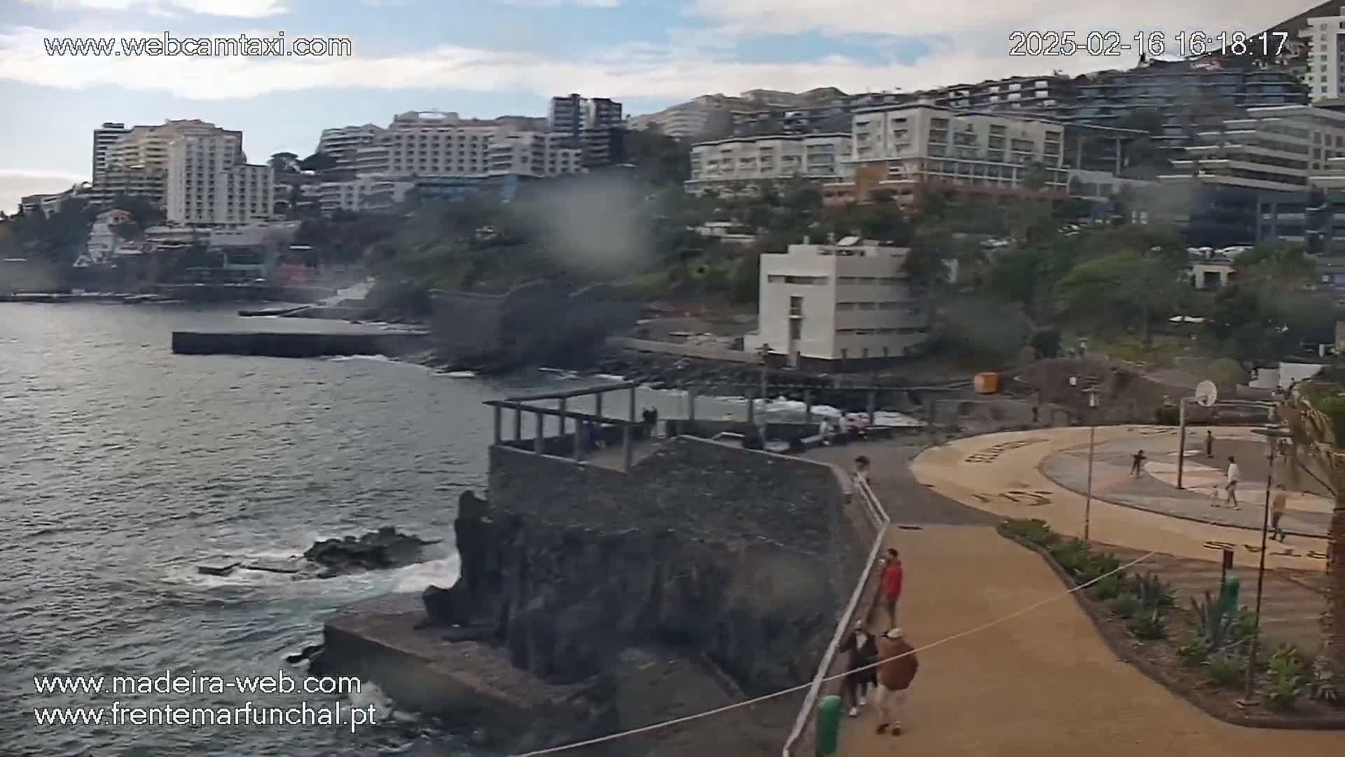 A coastal scene with overcast skies shows a rocky shoreline, buildings on a hillside, a paved area with people, and calm ocean water.