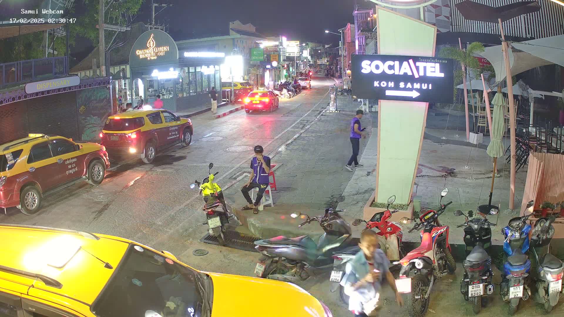 A nighttime street scene shows several parked taxis and motorbikes, with a few pedestrians and a person sitting on a curb, under clear skies.