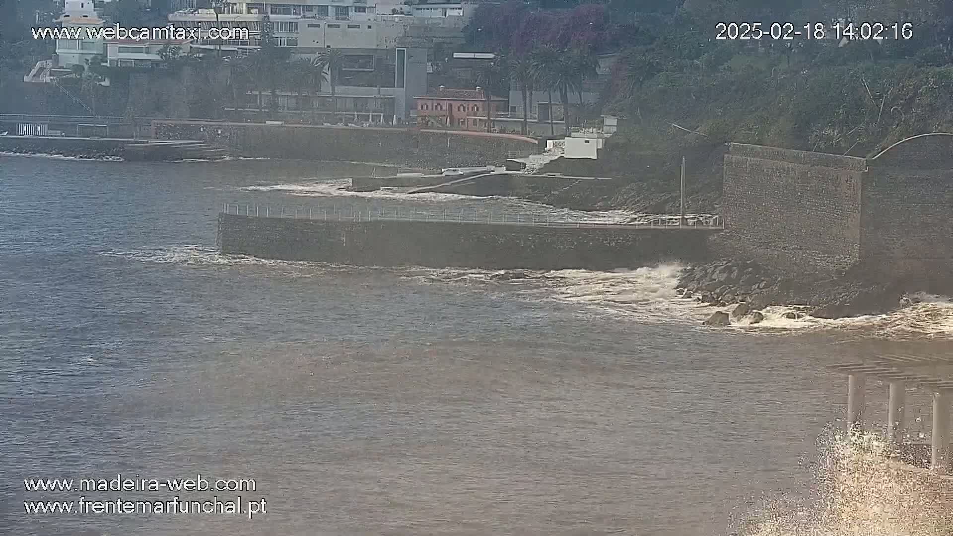 Waves crash against a seawall near a rocky shoreline with buildings visible in the background on an overcast day.