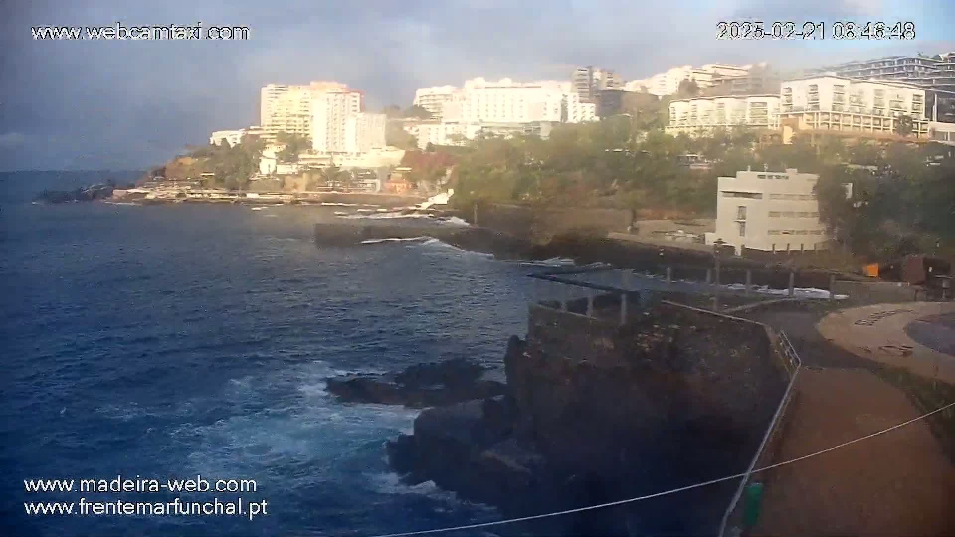 Ocean waves crash against dark volcanic rock formations near a coastline dotted with buildings under a partly cloudy sky.