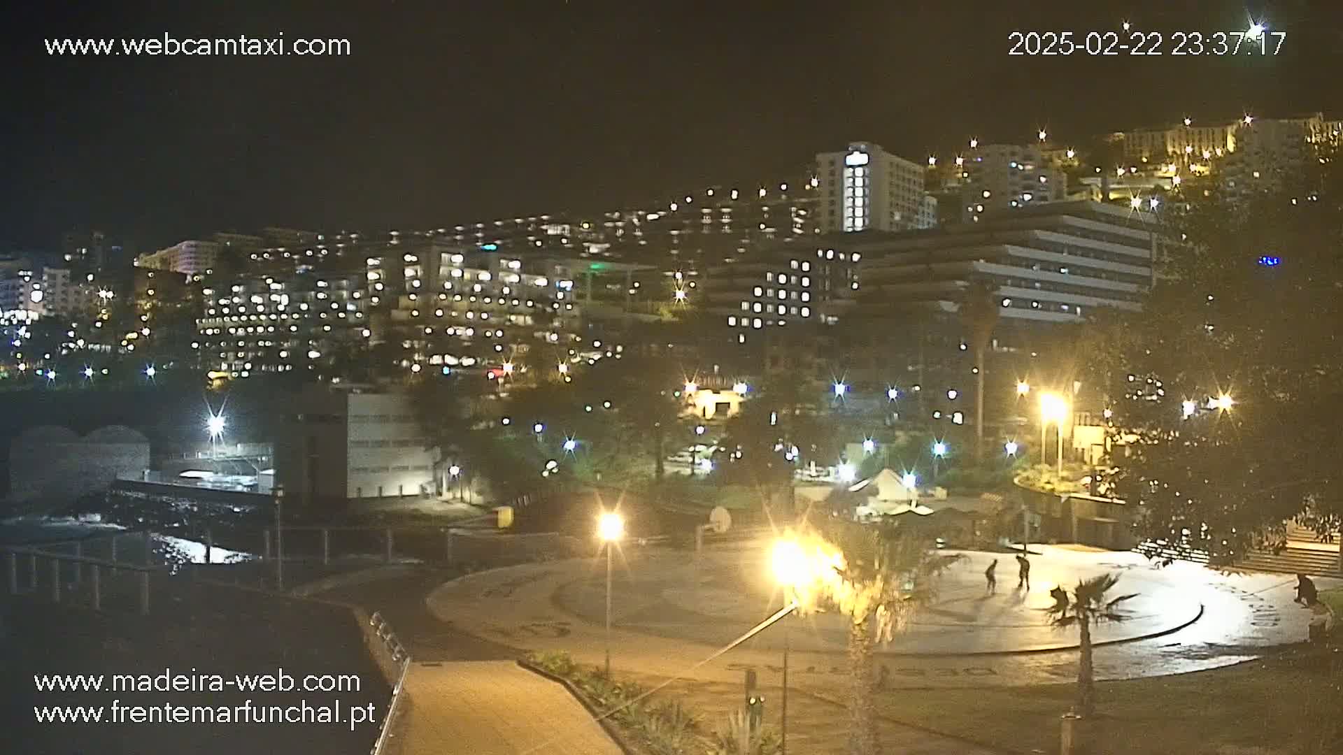 A nighttime view of a city illuminated by streetlights, featuring several multi-story buildings and a paved plaza with a few people.