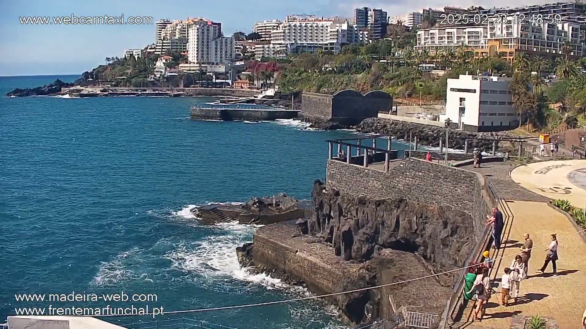 A coastal scene on a sunny day shows dark volcanic rock formations meeting a blue ocean with small waves, near buildings and people walking along a paved walkway.