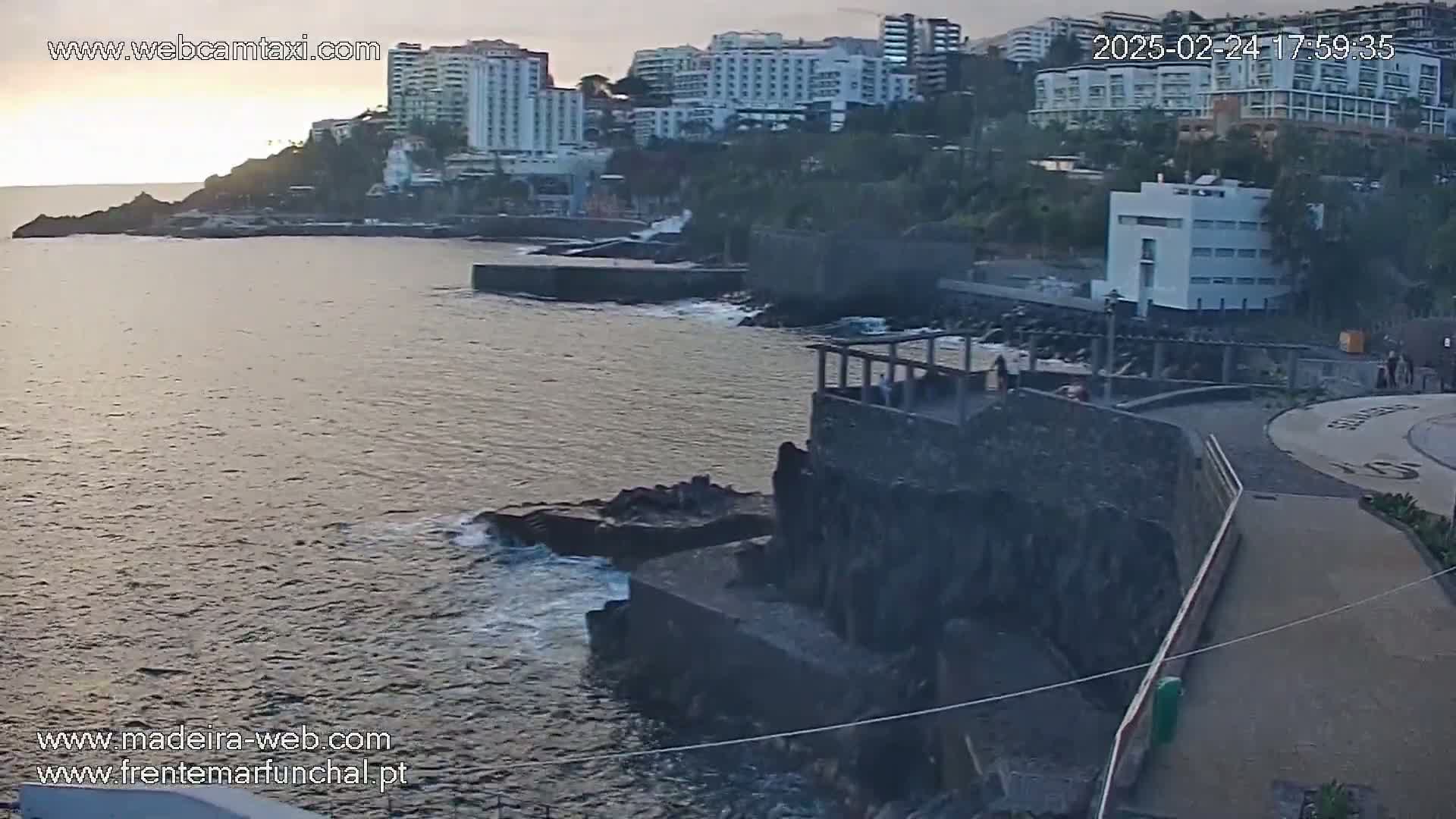 A calm ocean laps against dark volcanic rock formations near a coastal city with several hotels under a dusky sky.