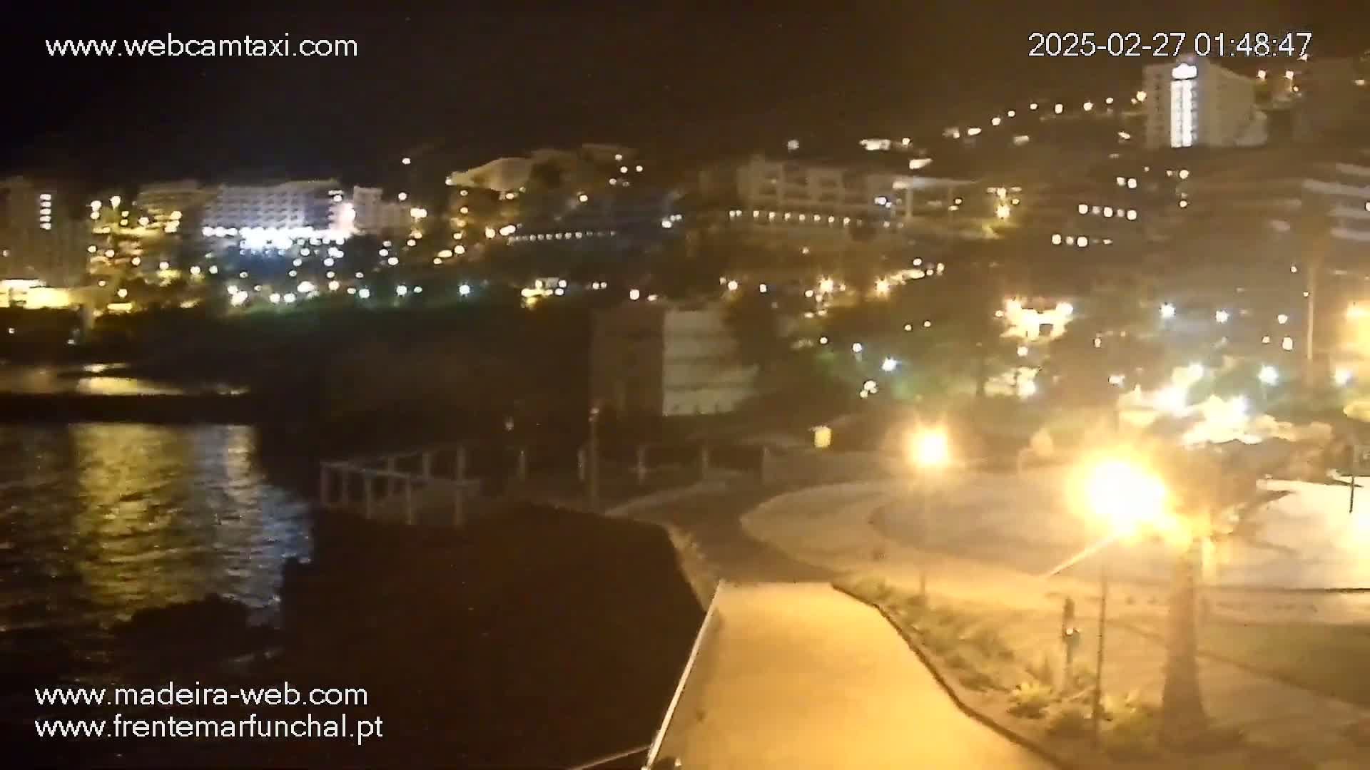 A nighttime coastal view shows a dark body of water, a paved walkway leading to a brightly lit area with buildings and palm trees on a hill.
