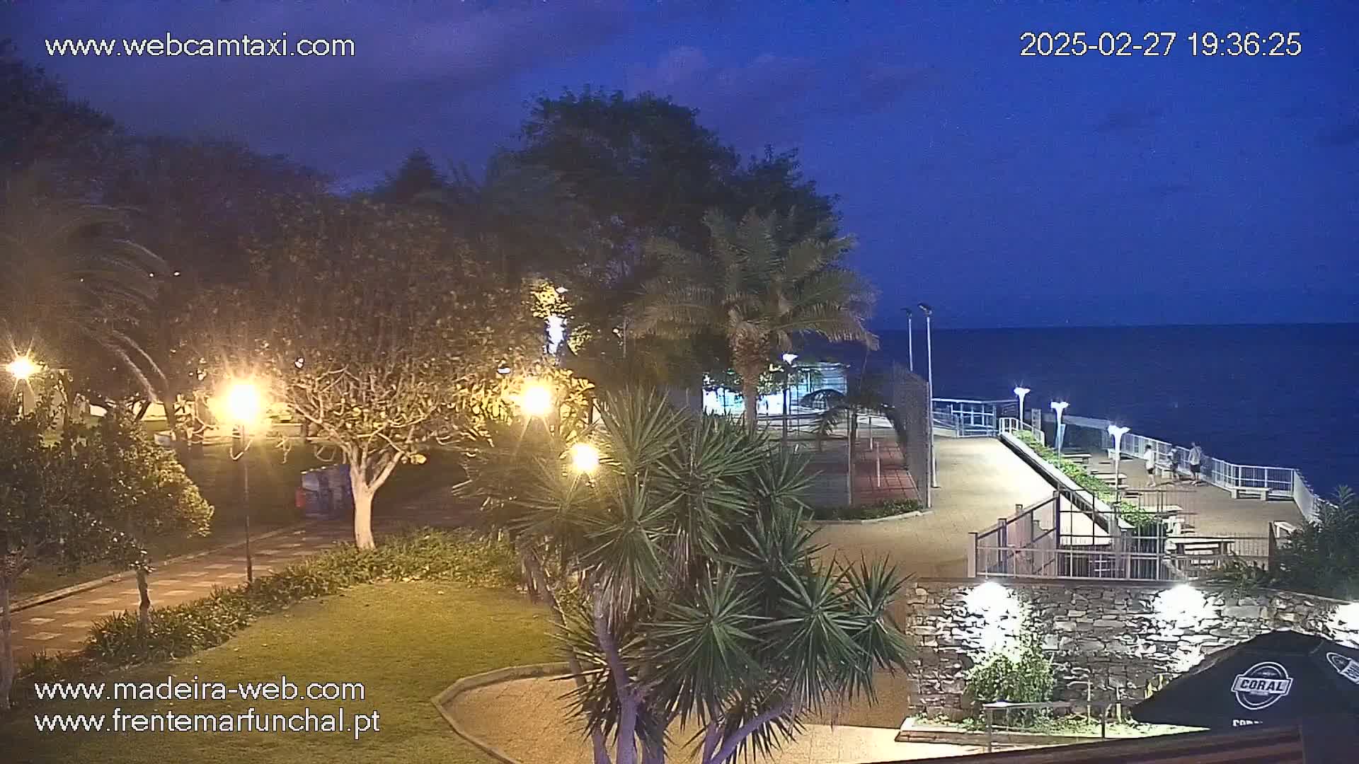 A nighttime view of a park-like area next to a paved waterfront walkway overlooking a dark ocean under a twilight sky.