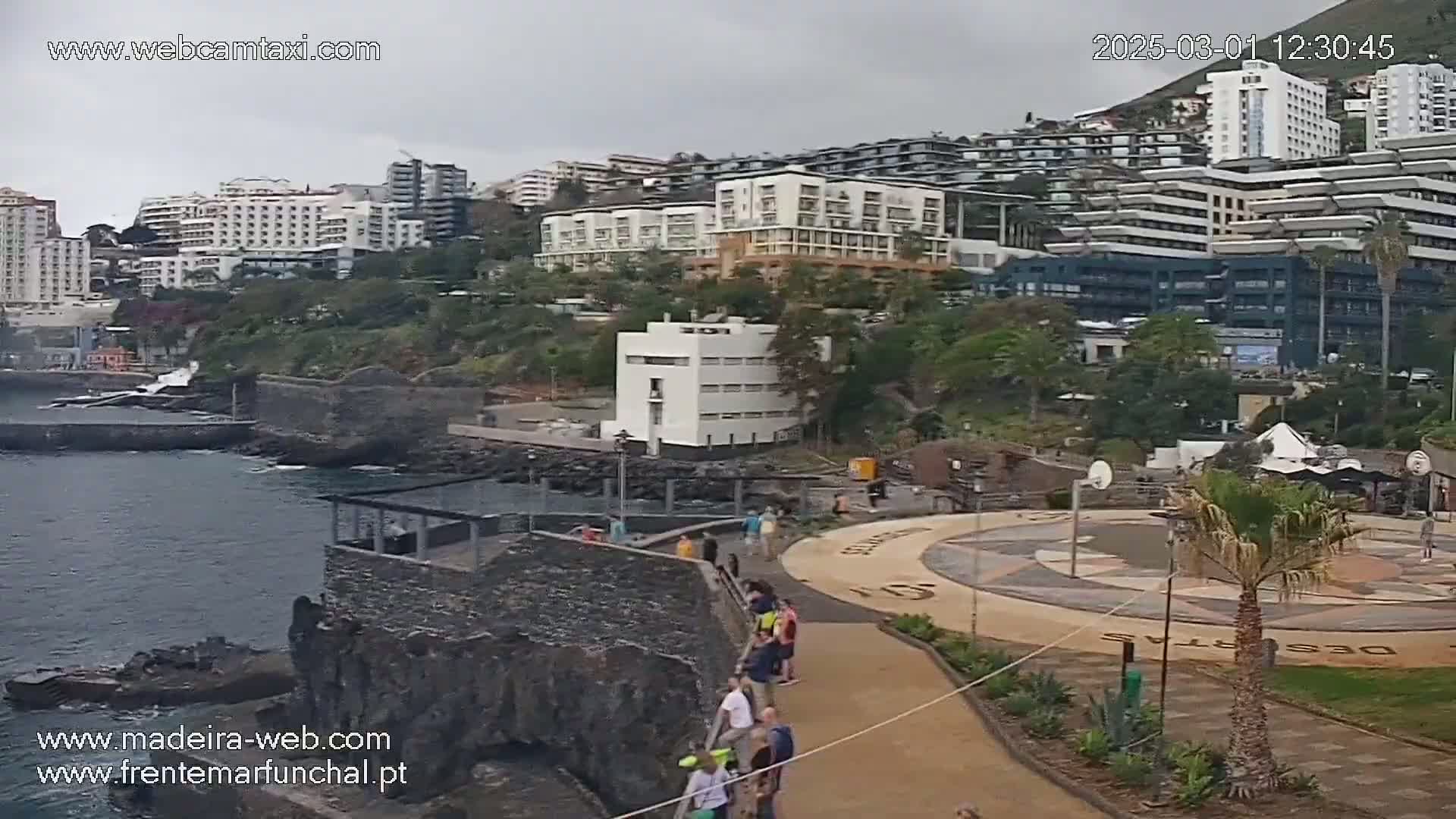 A coastal view on an overcast day shows several multi-story buildings overlooking a rocky shoreline with people walking along a paved path near a circular plaza.