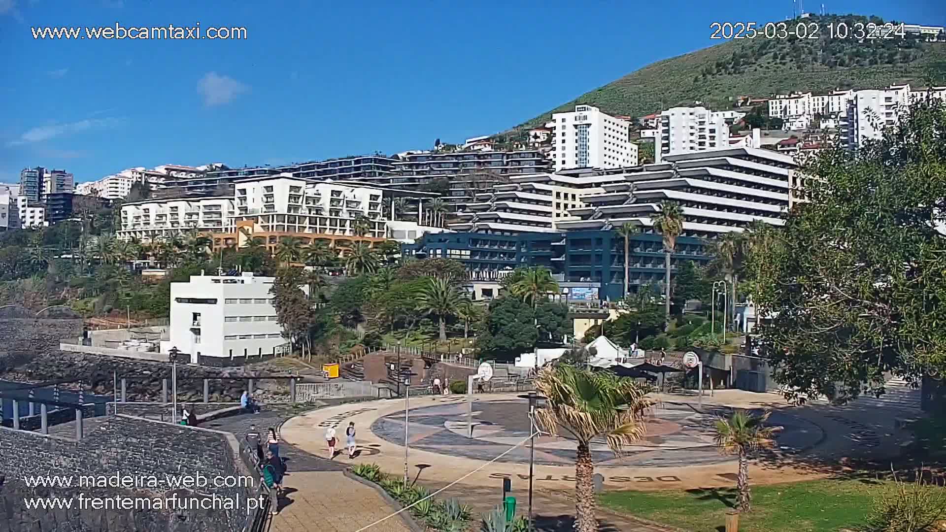 A sunny day overlooking a coastal city with numerous multi-story buildings, palm trees, and a paved plaza with people walking.
