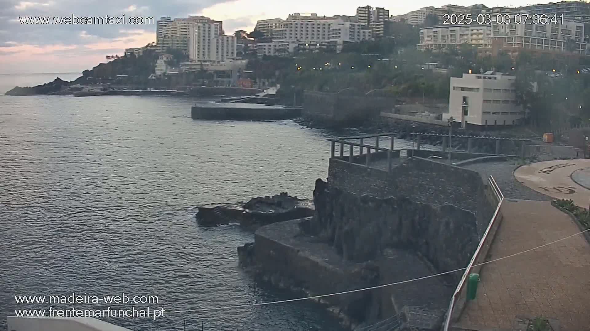 A coastal scene shows calm water next to a rocky shoreline with buildings and a paved walkway under an overcast sky.