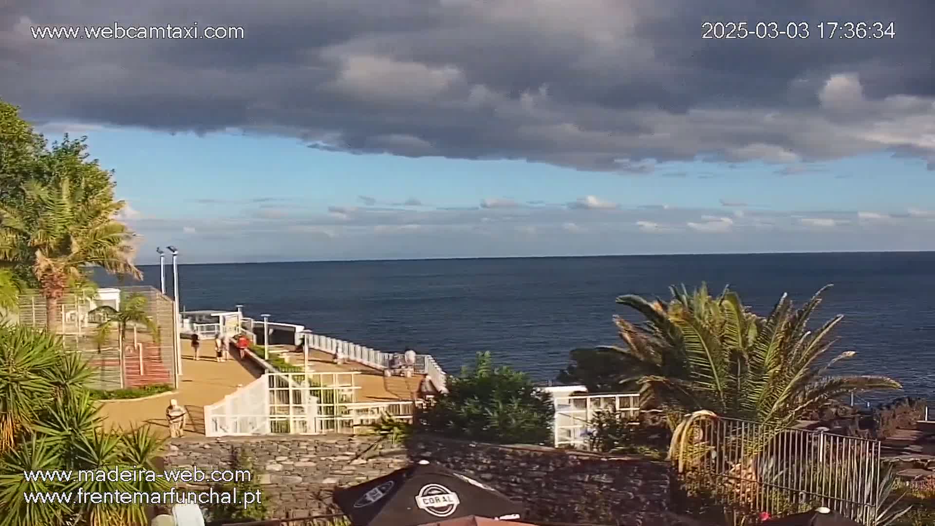 A partly cloudy day overlooks a calm ocean with a paved walkway and several people along the shoreline, surrounded by palm trees and other tropical vegetation.