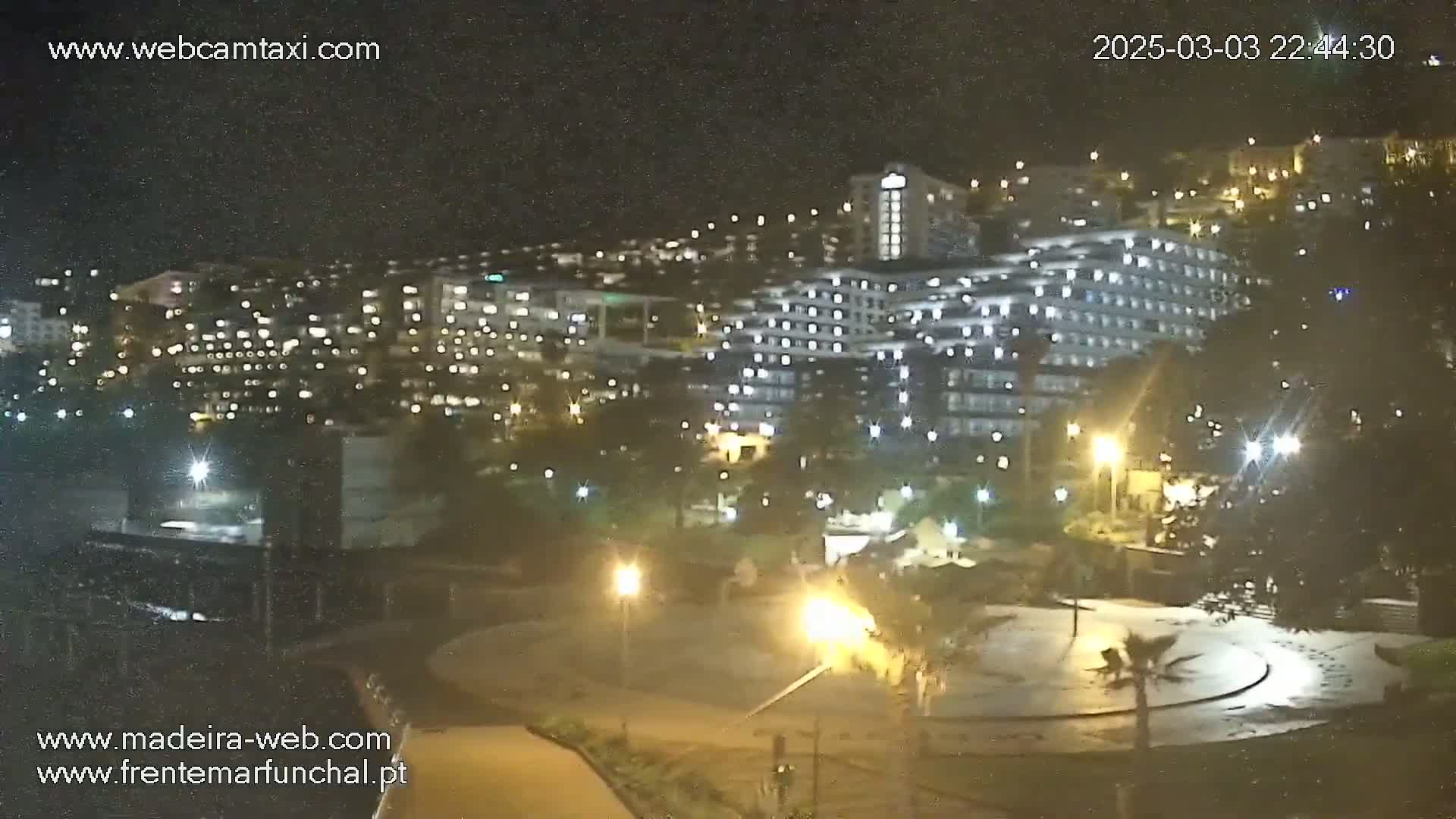 A nighttime view of a city with numerous illuminated buildings and a plaza, under a slightly hazy sky.