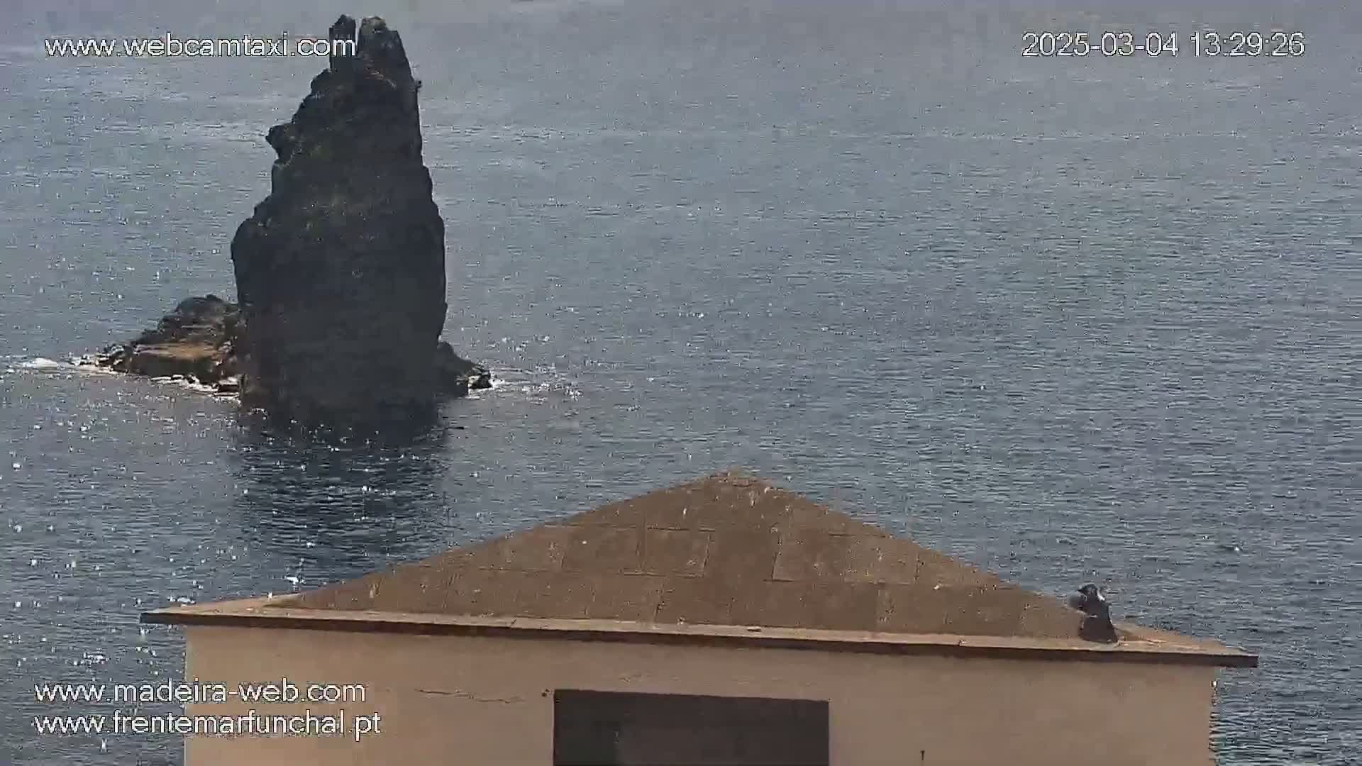 A dark, tall rock formation sits in a body of relatively calm water, partially visible behind a tan, triangular-roofed structure.