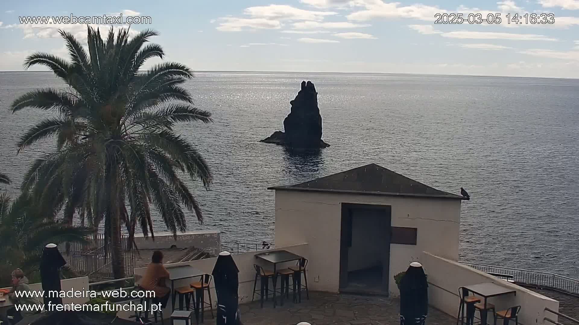 A seaside patio with tables and chairs overlooks a dark rock formation in the ocean under partly cloudy skies.