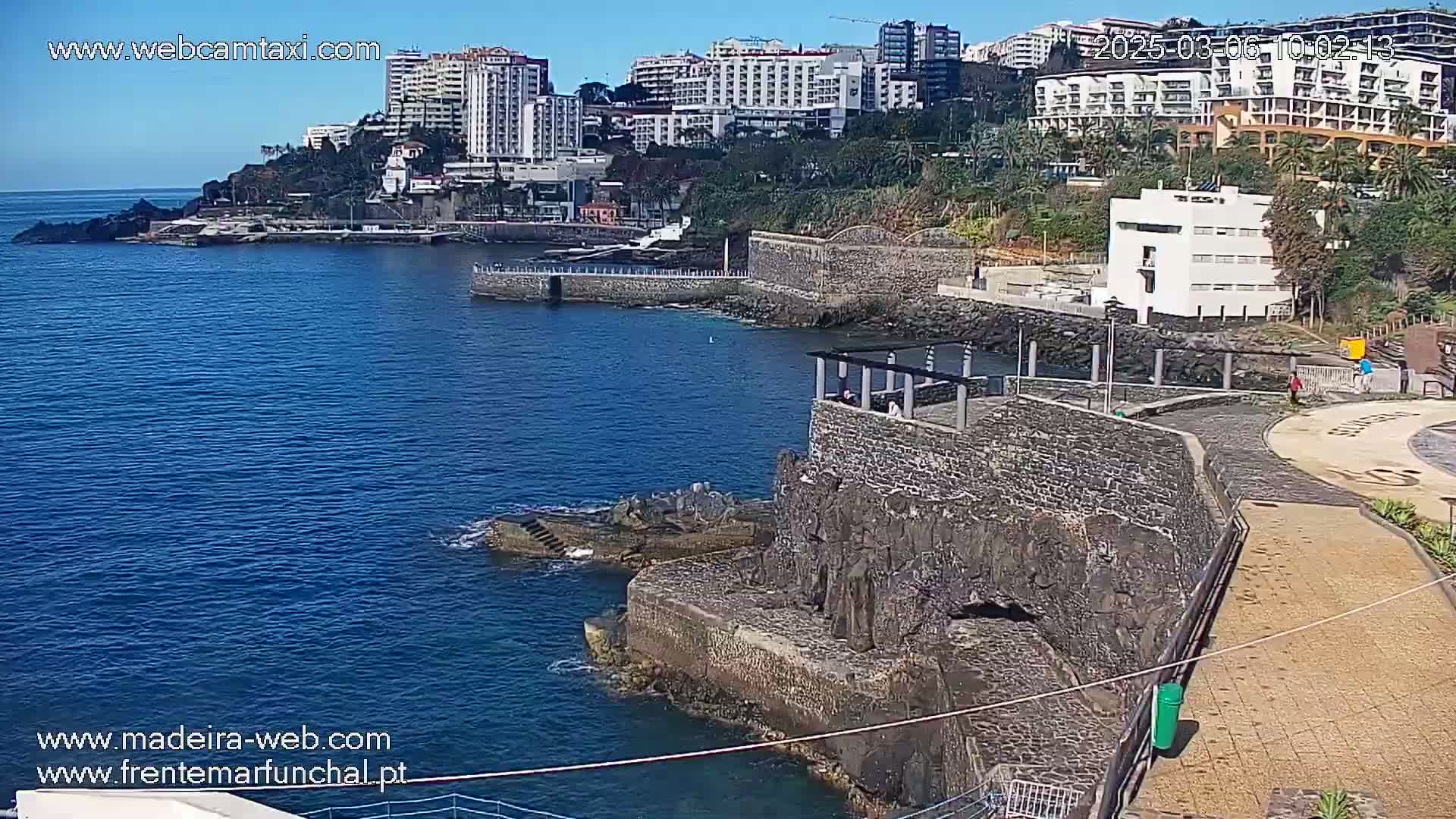 A rocky shoreline with a stone structure and calm blue water is seen under a sunny sky, backed by multi-story buildings and lush vegetation.