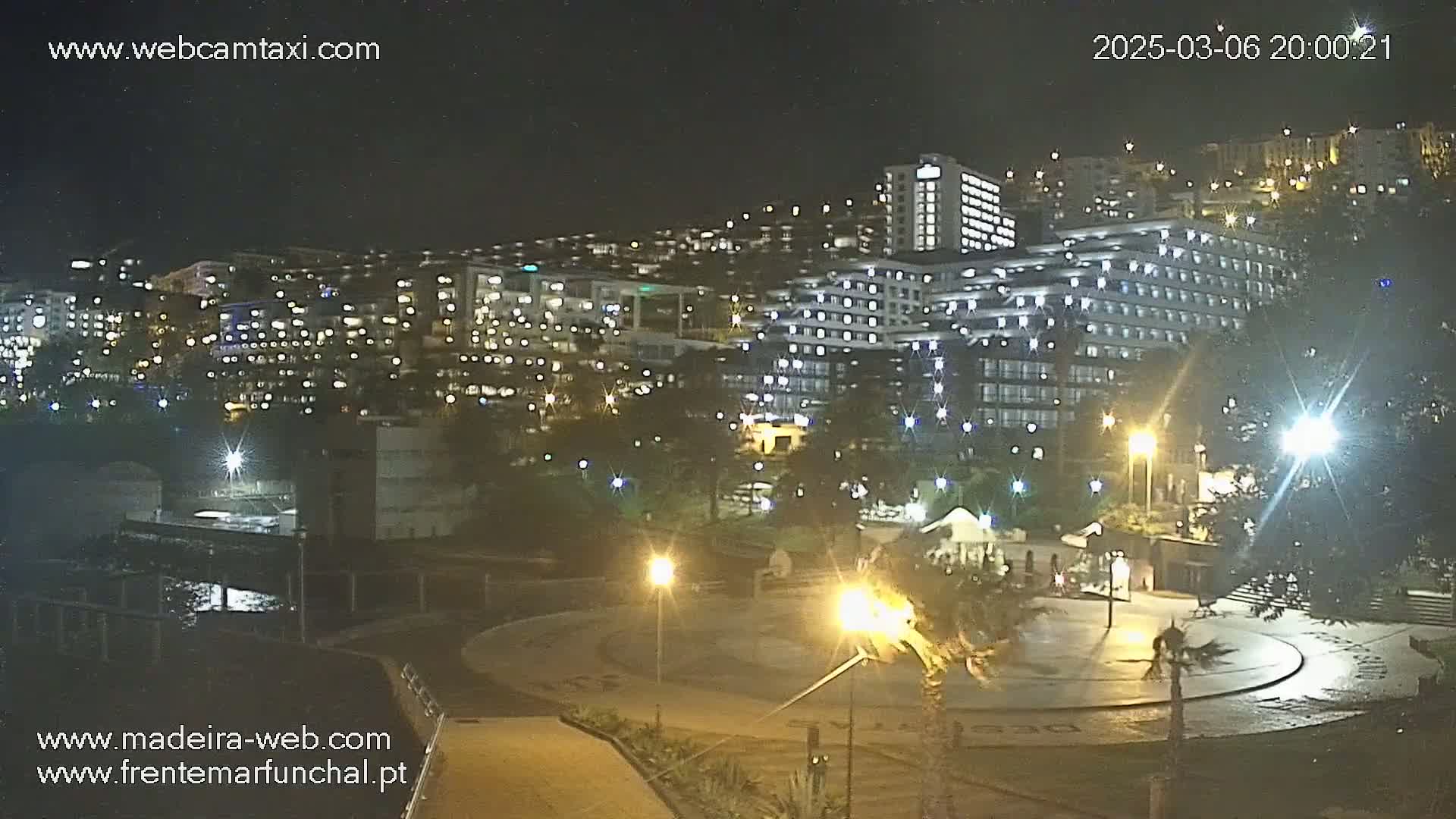 A brightly lit, multi-story hotel complex is seen at night, surrounded by trees and a paved plaza with a few people visible.